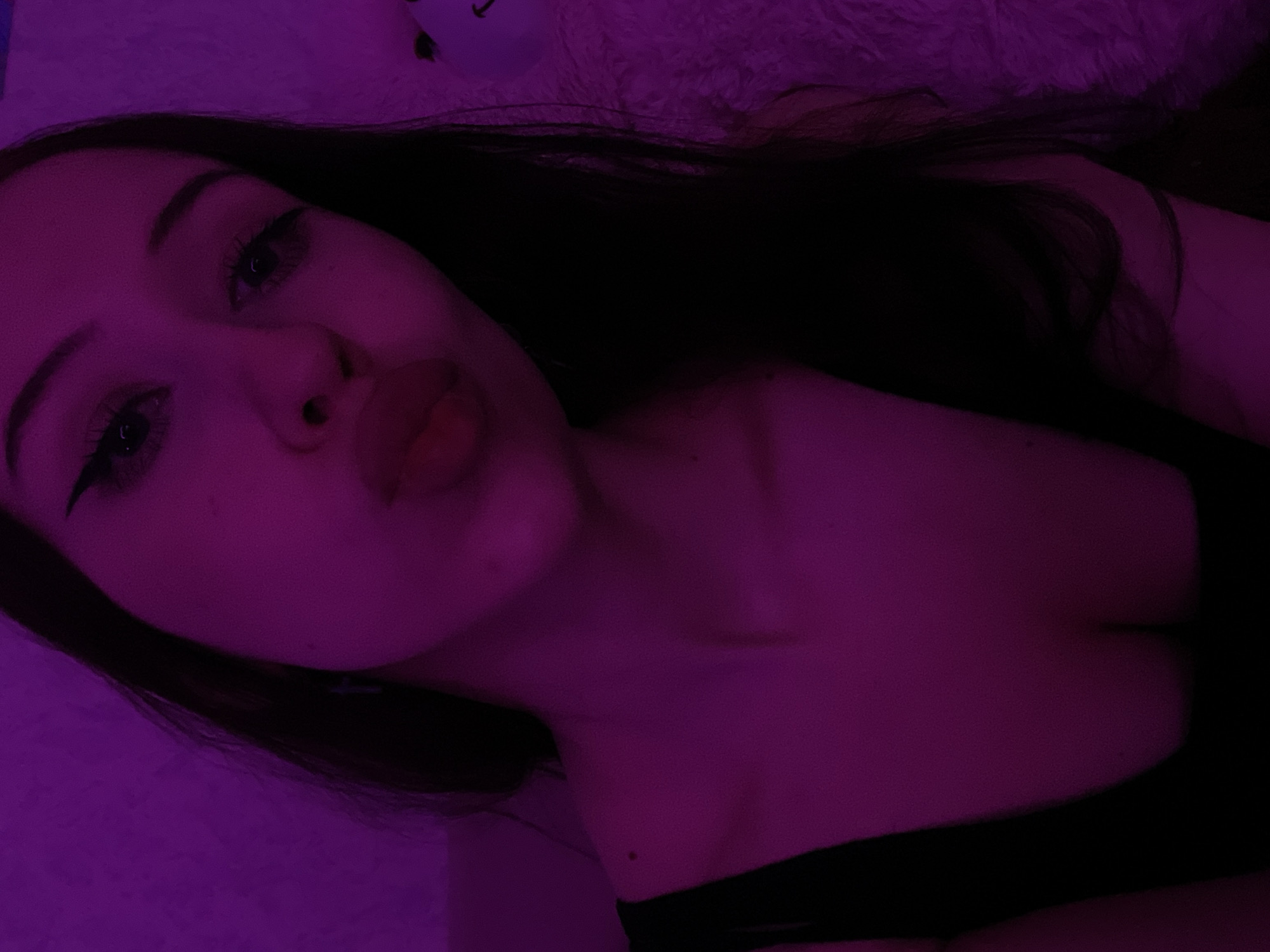 A young woman with dark hair, wearing a black top, is illuminated by purple light. She is looking towards the camera with her lips puckered, as if for a kiss. Her eyes are lined with eyeliner, and her eyebrows are arched. The background is a solid purple color, possibly a wall. There is a fluffy white object on the right side of the image, partially obscured by the woman's hair. The overall impression is a selfie taken in a dimly lit room with colored lighting. The image does not provide any specific clues about its location in Votkinsk, Russia, or any other geographical context.