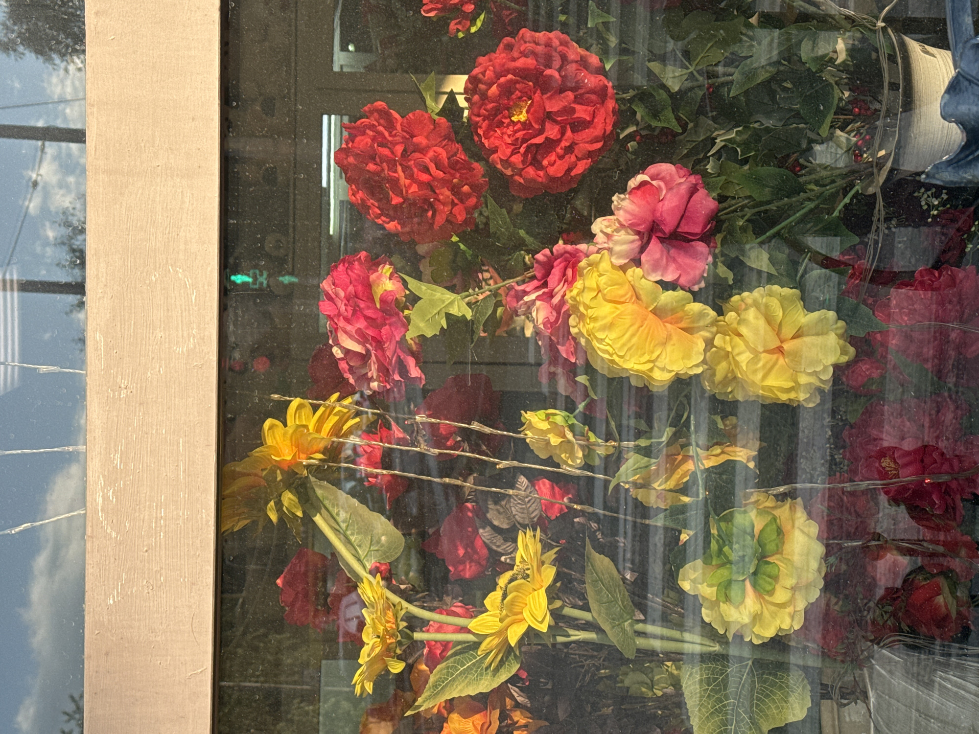 The image shows a display of artificial flowers in a window, with reflections of the sky and trees visible on the glass. The flowers are a mix of vibrant colors, including yellow sunflowers, red roses, and pink peonies. The display appears to be in a shop window, possibly a florist or a gift shop. The overall impression is one of a cheerful and colorful arrangement. There are no people visible in the image, and no specific actions or interactions are depicted. The environment appears to be outdoors, given the reflections of the sky and trees. There are no indications of the location in Saint Petersburg, Russia.