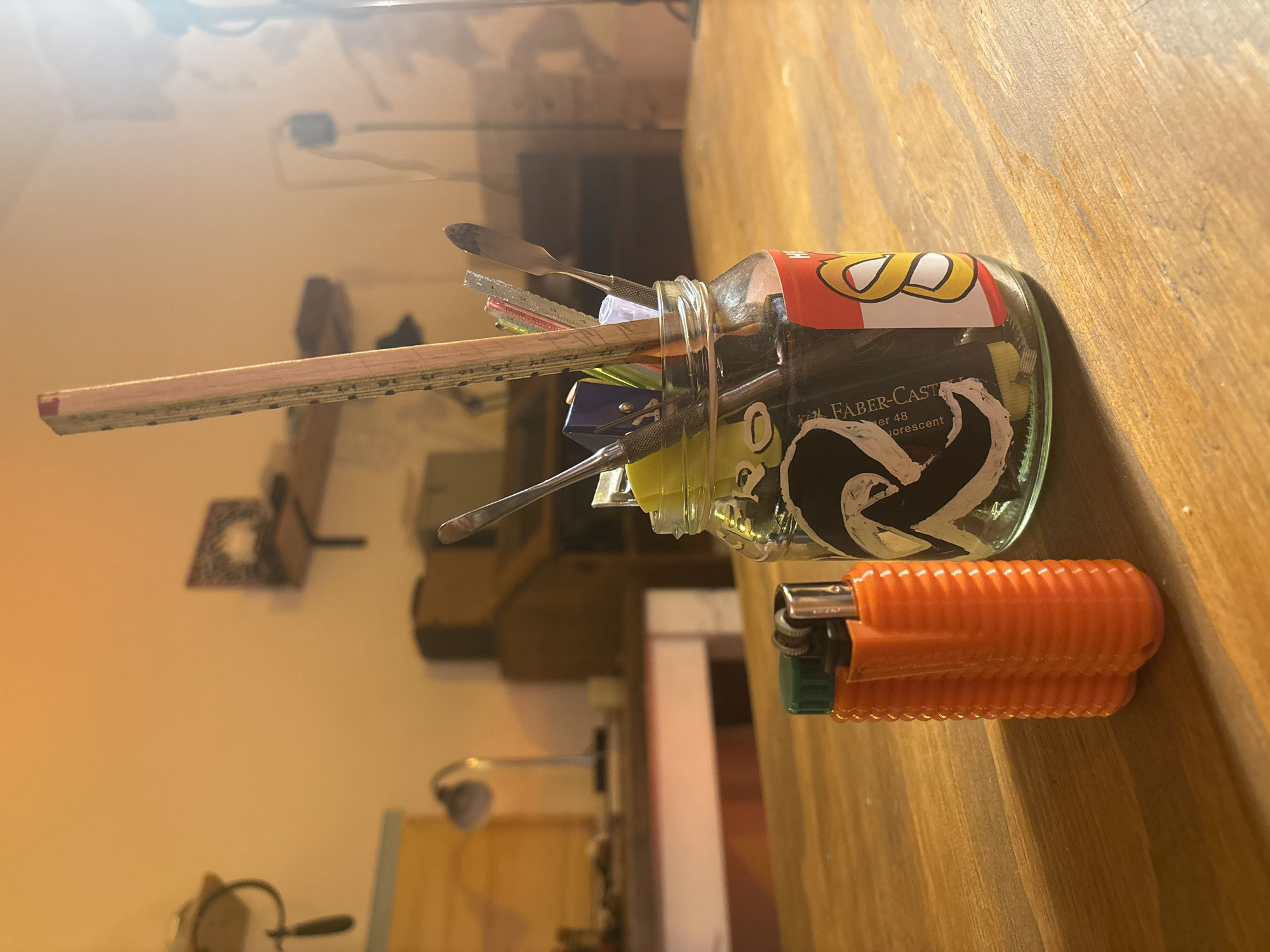 A glass jar filled with various items sits on a wooden surface, alongside an orange lighter. The jar contains a pencil, several small tools, and assorted objects. The jar has labels on it, including "Faber-Castell." The wooden surface appears to be a table. The background includes indistinct objects and lighting fixtures. There's no indication of the location being in Buenos Aires, Argentina.