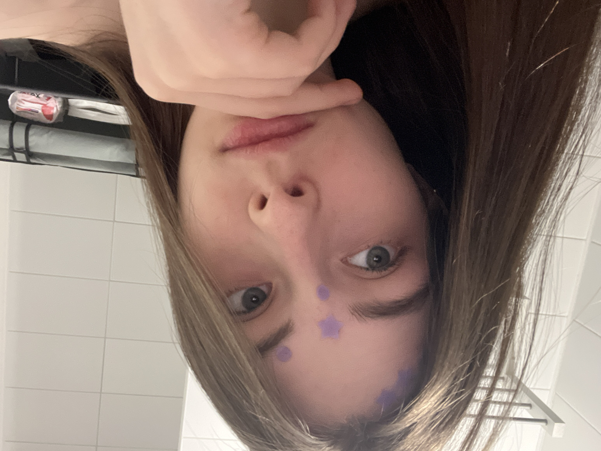 A close-up selfie-style image shows a young person, likely female, with long, light brown hair and fair skin, looking directly at the camera. The photo is oriented 90 degrees counter-clockwise, so the subject appears upside down within the frame. She has large, light-colored eyes and a neutral, slightly curious expression. Her left hand is positioned with fingers partly covering her mouth and chin. Several small purple shapes, including circular dots and a star, are applied to her forehead and left cheek, resembling face paint or stickers.
The background suggests an indoor setting, likely a bathroom, with white rectangular wall tiles. In the upper left background (if the image were correctly rotated), a small red and white package is visible on a shelf or ledge. Partially visible text on this package appears to be "ATAX" or "VATRA." Below it, a white cylindrical object and a flat white surface can be seen. On the right side of the background, a simple metallic rack or rail is present. The overall scene is a personal snapshot. The implied location is Saint Petersburg, Russia.