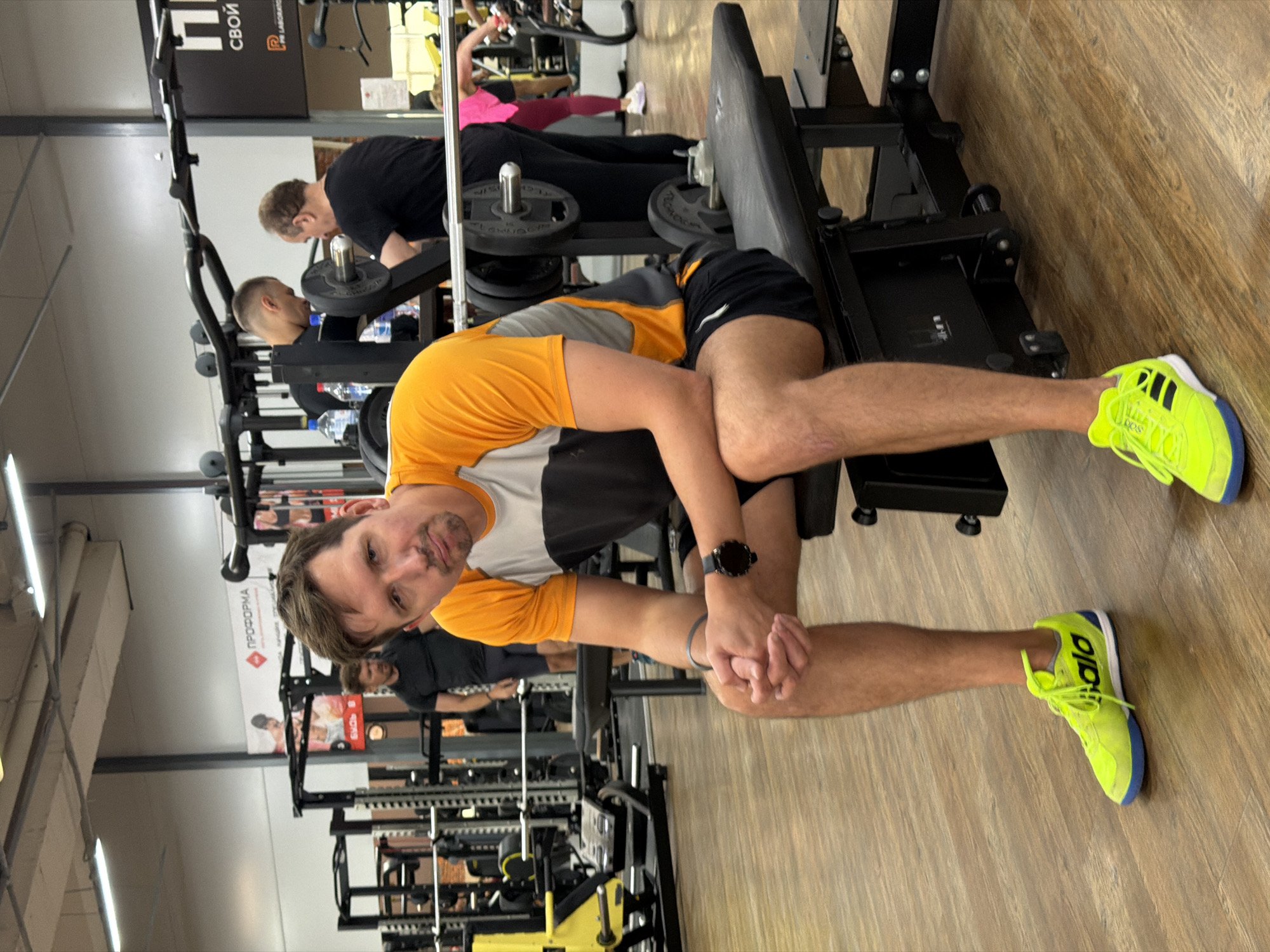 The image captures a candid moment in a gym located in Chaikovsky, Russia, with the main subject looking directly at the camera. A man with short brown hair, wearing an orange and grey t-shirt, black shorts, and striking neon yellow Adidas sneakers, is seated on a gym bench, resting his hands clasped between his legs. A black smartwatch is visible on his left wrist. He has a neutral to slightly smiling expression.
In the background, other individuals are engaged in their workouts. To the left, another man with short hair is bent over a piece of gym equipment, possibly adjusting weights or performing an exercise. Further back and to the right, a person in a bright pink top and dark leggings is seen on another machine. Various pieces of weightlifting equipment, including racks, benches, and weighted plates, are scattered throughout the gym. Several water bottles are also visible near the equipment.
The gym features a light wooden floor and bright artificial lighting from the ceiling. A prominent black sign in the background displays the Russian word "СВОЙ" (Svoy, meaning "own" or "one's own") and "PR LABORATORY." Another red sign partially visible further back reads "ПРОФОРМА" (Proforma). The overall atmosphere appears active and typical of a modern fitness center.