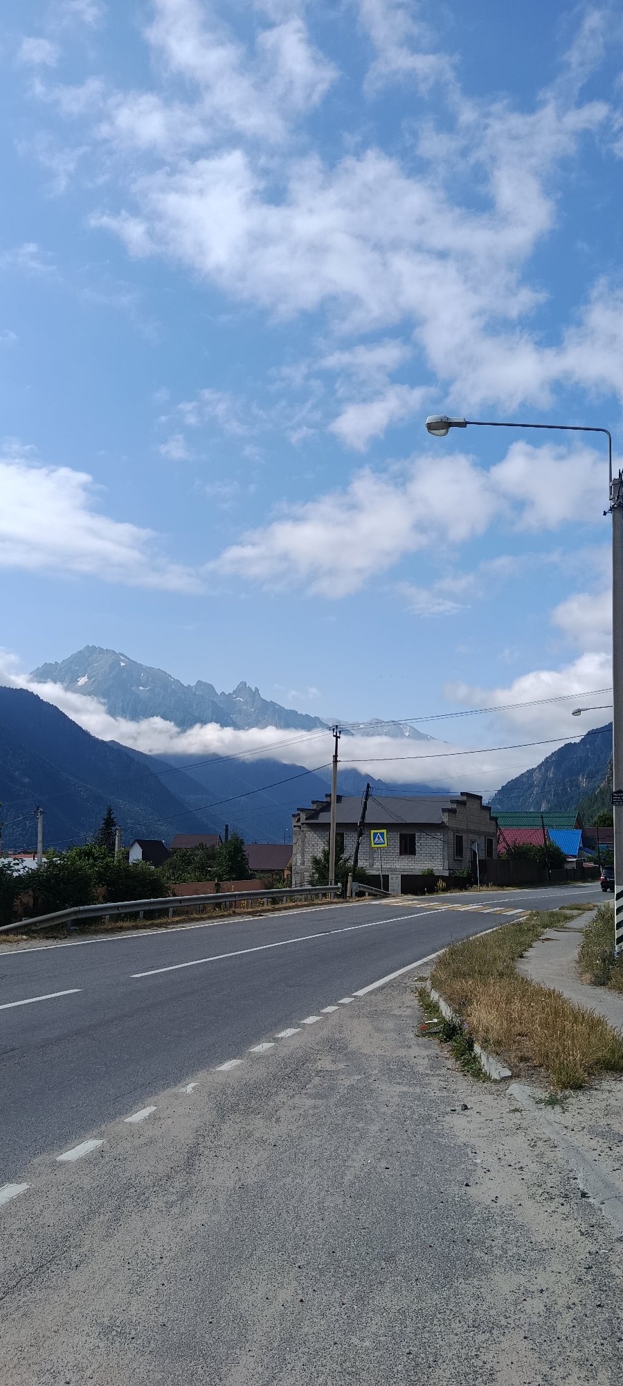 The image shows a view of a road in Teberda, Russia, with mountains in the background. The road is paved and has white lines marking the lanes. There are several houses and trees visible on the sides of the road, as well as a lamppost and a pedestrian crossing sign. The mountains are tall and majestic, with some of them covered in clouds. The sky is blue with scattered white clouds.