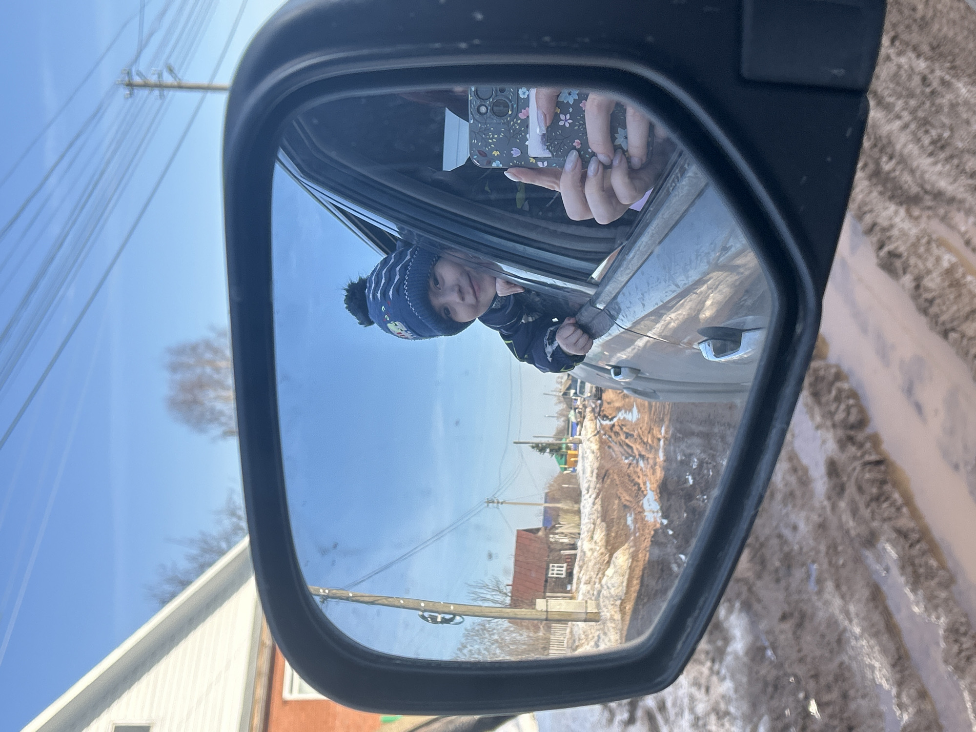 The image shows the reflection in a car's side mirror. A child wearing a blue hat and jacket is looking out the car window, partially obscured by the window frame. Another person is holding a phone with a floral patterned case, apparently taking a photo. The reflection also shows a roadside scene, including houses, utility poles, and muddy ground. This scene is likely in Фоки, Russia.