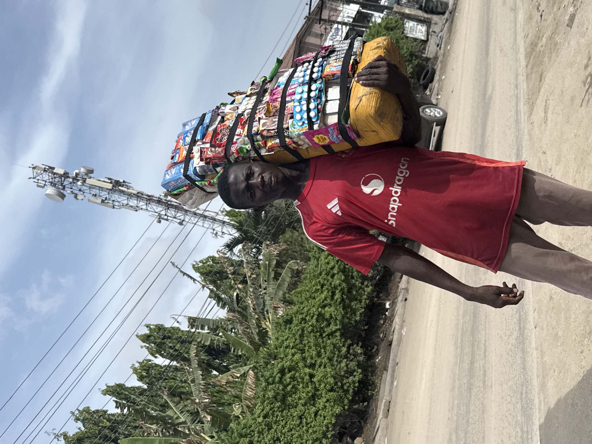 A young Black man, likely a street vendor, is captured in a vibrant outdoor scene in Ado, Nigeria, carrying a substantial load of merchandise on his head. He stands on a dirt road, looking directly towards the viewer with a neutral expression. He is wearing a red short-sleeved t-shirt featuring the white "S" logo and "Snapdragon" text, with a small Adidas logo beneath. His lower body is clad in dark trousers or shorts.
The elaborate bundle on his head consists of numerous packaged goods, including snacks, candies like "Mentos" and "Chupa Chups," and blister packs that appear to contain tablets or capsules. The items are neatly arranged and secured with black straps, forming a tall, rectangular stack. His hands are raised to support a yellow wrapped parcel that rests on his shoulders, acting as a base for the main load.
The setting appears to be an urban or semi-urban area, characterized by a unpaved road with a curb, lush green foliage including identifiable banana trees, and various other trees and bushes lining the path. In the left background, a tall metal communication tower is prominent against a bright sky with scattered clouds, indicating clear daytime conditions. Power lines crisscross the upper left portion of the frame. A white car is partially visible on the right side of the road in the background.