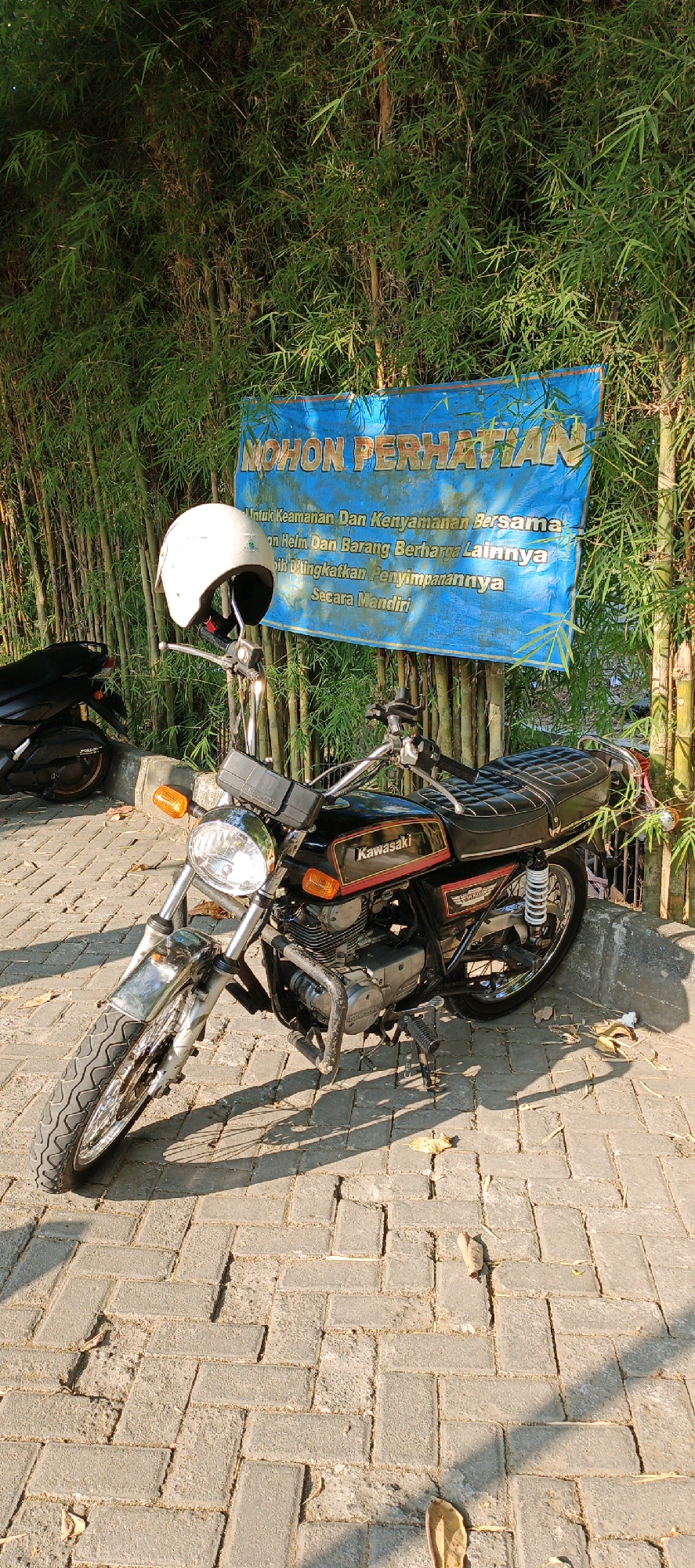 This image depicts a Kawasaki motorcycle parked outdoors on a paved surface in Trihanggo, Indonesia. The motorcycle is black with red and gold accents and a white helmet is placed on its handlebars. Behind the motorcycle, a bamboo fence forms a backdrop, with a blue banner displaying text in Indonesian that translates to "ATTENTION". The banner appears to be a notice about safety and valuable item storage. The sun is casting shadows, indicating it is likely daytime. The ground is covered with interlocking brick pavers, and a few fallen leaves are scattered around. In the background, another motorcycle is partially visible.