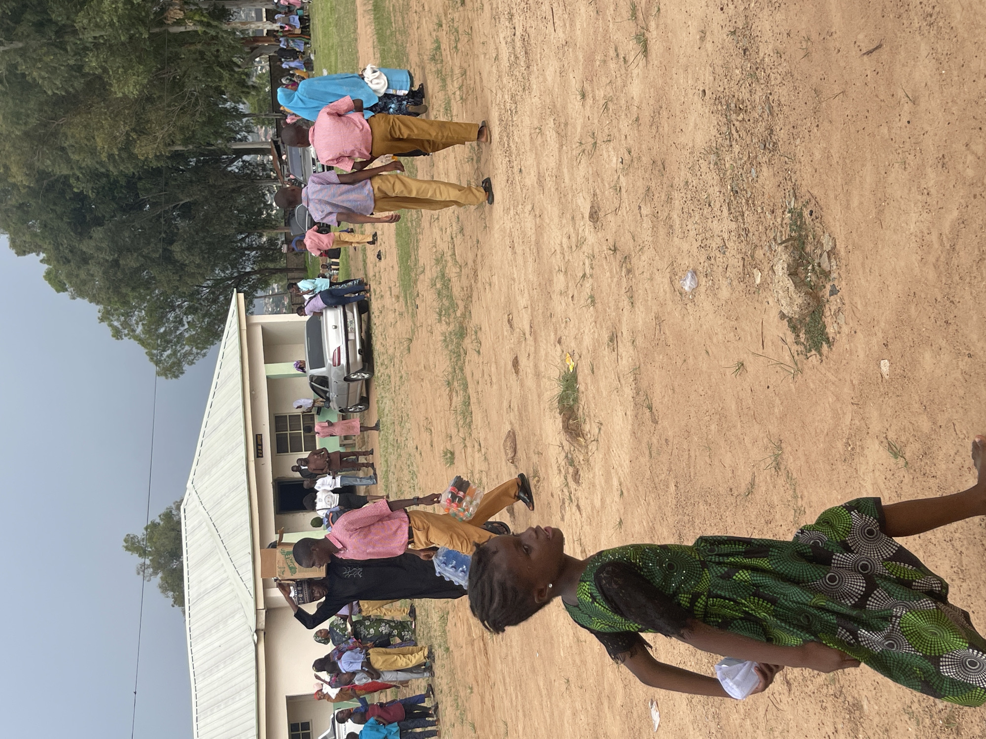 In Unguwar Saya, Nigeria, a group of people are gathered outside a light-colored building with a white corrugated metal roof. The building appears to be a public facility, with signage indicating "IRS REA OFFICE" above the entrance. Several individuals are walking and standing in the foreground and midground. A young girl in a green and black patterned dress is walking towards the left in the foreground, holding a white object. In the midground, several men and boys are dressed in traditional attire and modern clothing, some carrying bags and boxes. A man in a black traditional outfit is carrying a large cardboard box on his head. To his right, a man in a pink shirt and tan pants is walking with two plastic bags containing bottles. Several cars are parked in the background, including a silver sedan. The ground is dirt with sparse vegetation and scattered debris. The sky is overcast. The scene suggests a communal gathering or distribution event.