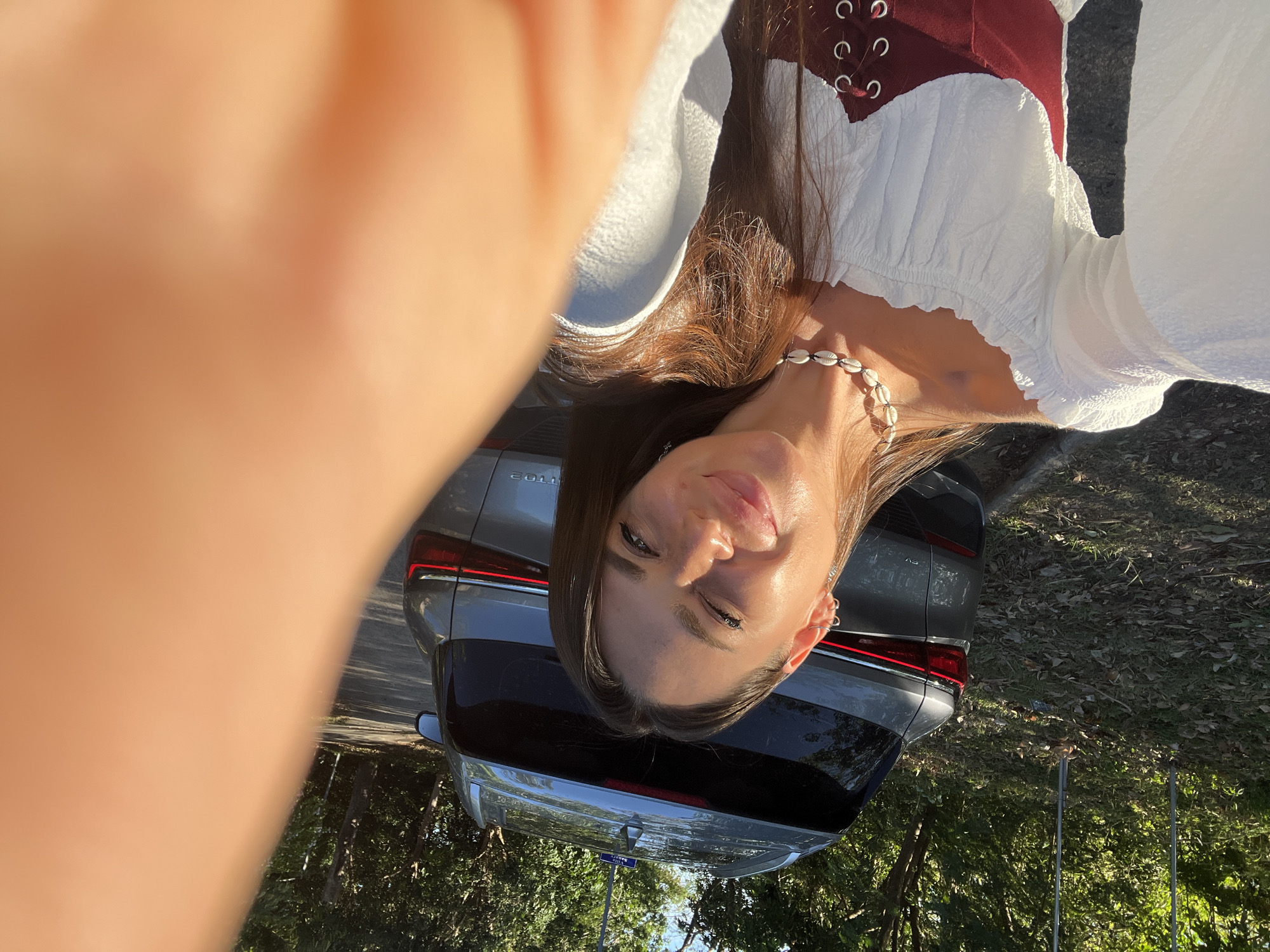 A young woman with long brown hair is taking a selfie outdoors. She is wearing a white off-the-shoulder top with a dark red corset over it and a seashell necklace. She is smiling and her left eye is partially closed. Behind her, a gray SUV is parked on a paved road. The environment is sunny with trees and foliage in the background. There is a sign on the roof of the SUV.