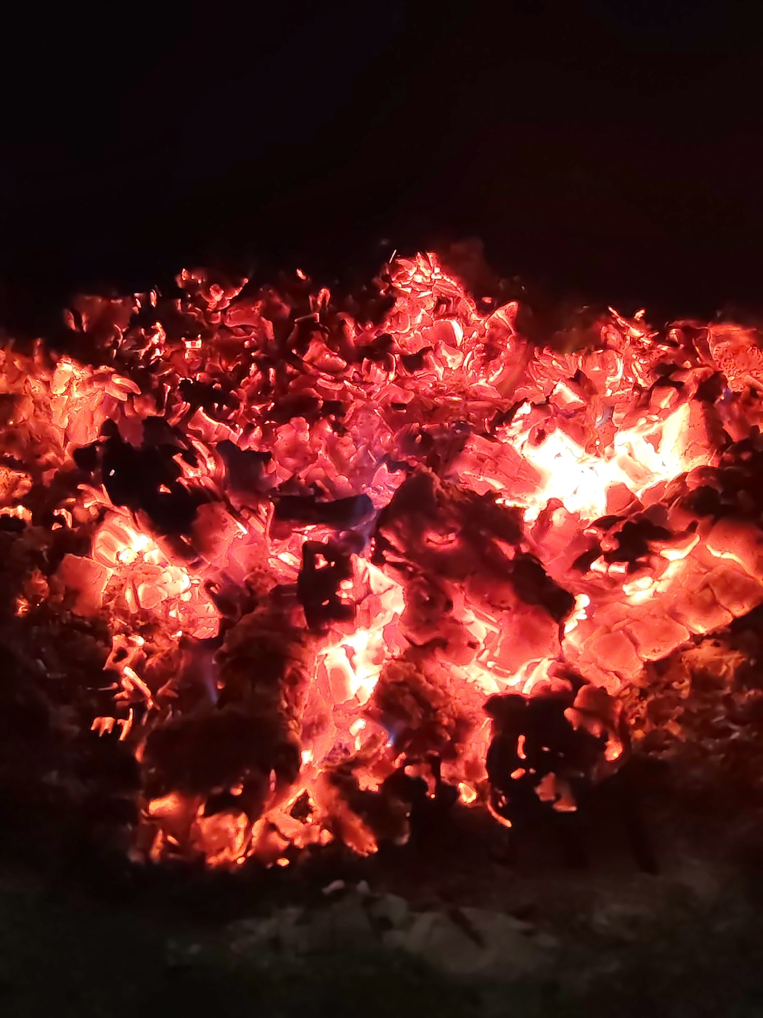 The image shows a bed of glowing embers in the dark. The embers are primarily red and orange, with some brighter white-hot spots. The background is a solid dark black. The location is unknown.