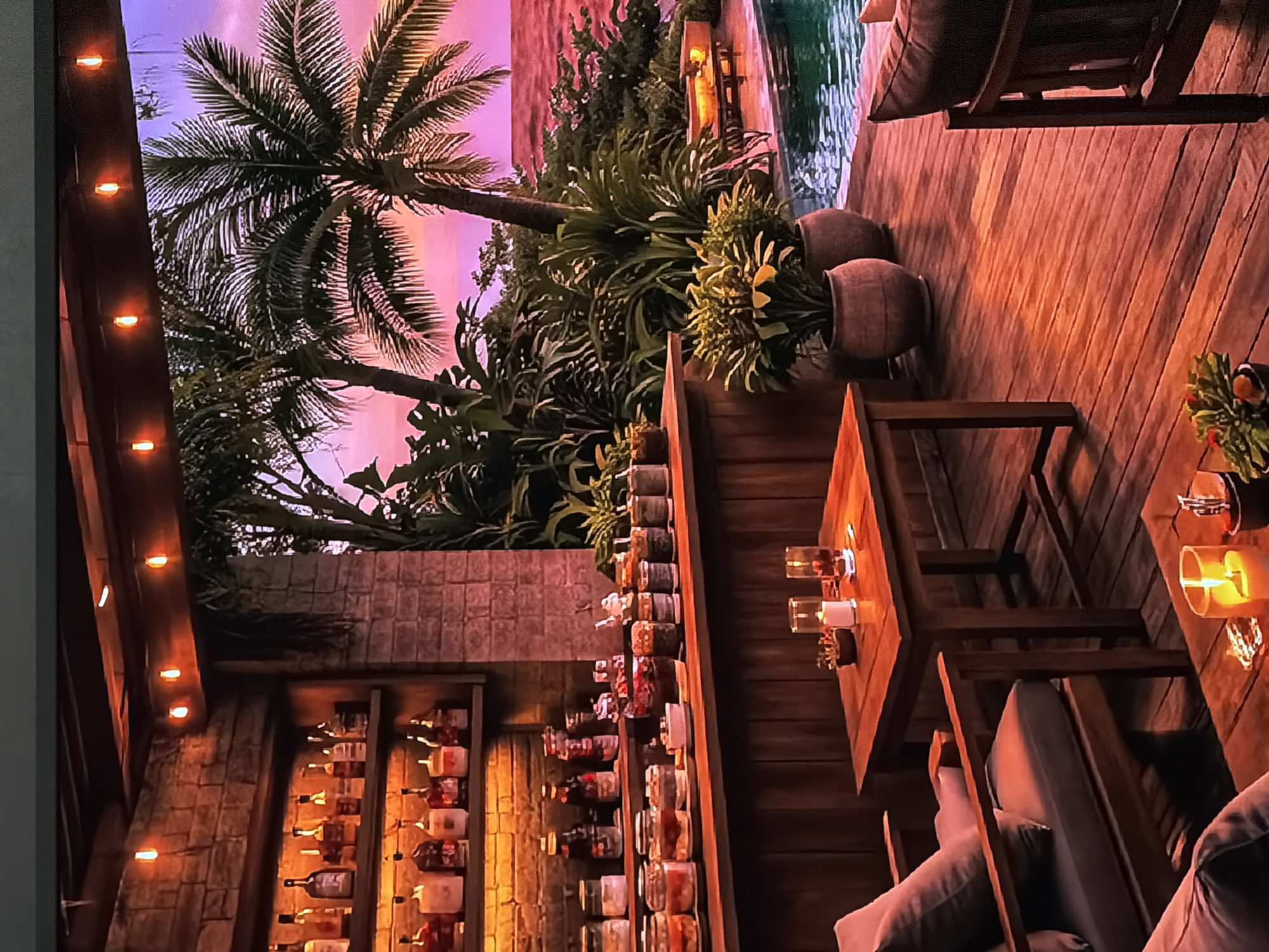The image displays a luxurious and inviting outdoor bar and lounge area, likely captured at dusk or in the early evening, in Sofiivska Borshchahivka, Ukraine. The scene exudes a warm, tropical ambiance, suggesting a specially designed entertainment space.
To the right, a well-maintained wooden deck stretches out, featuring several sturdy wooden tables and chairs with light-colored cushions, set for guests. Multiple lit candles in glass holders are placed on the tables, casting a soft, warm glow that contributes significantly to the cozy atmosphere. Near the deck's edge, two large, earthy-toned pots hold lush green plants, adding to the natural decor. A serene swimming pool is visible adjacent to the deck, reflecting the ambient light.
The background is rich with tropical foliage, including tall palm trees and dense, varied greenery, creating a vibrant, resort-like environment. The sky above transitions from a soft pink to purple hue, characteristic of twilight.
On the left side of the scene, an illuminated outdoor bar stands against a textured stone or brick wall. Its shelves are well-stocked with numerous bottles, likely spirits and wines, and rows of jars possibly containing bar ingredients or garnishes. Above the bar area, a wooden beam or pergola is adorned with warm string lights, further enhancing the festive and intimate evening mood.
The entire setting is meticulously arranged to evoke a sense of relaxation and luxury, ideal for evening entertainment or quiet enjoyment. No people are visible, allowing the focus to remain on the beautifully crafted environment and its tranquil yet inviting atmosphere. There is no visible text in the image.