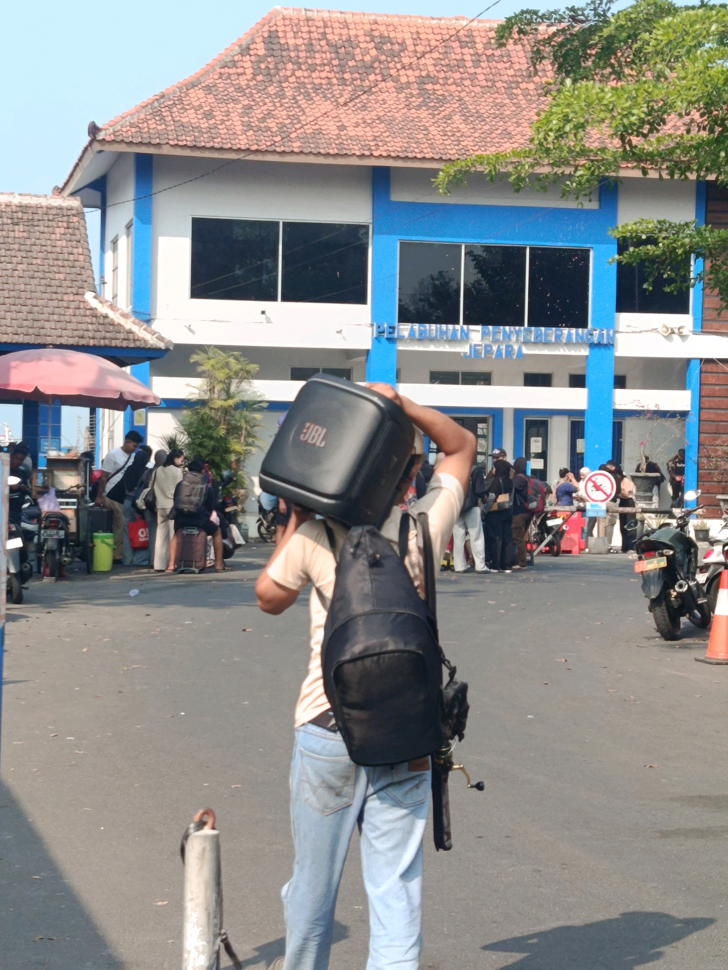 The image depicts a scene outside a building in Jepara, Indonesia, likely a port or ferry terminal, as indicated by the text on the building which reads "PELABUHAN PENYEBERANGAN JEPARA". In the foreground, a person is carrying a large black JBL speaker on their right shoulder, with a black backpack also on their back. This person is wearing light blue jeans and a light-colored t-shirt. Behind them, a group of people are gathered near a structure with a red umbrella, and several motorcycles are parked. The building in the background has a red tiled roof and white walls with blue accents around the windows and along the structure's edges. The ground is paved and appears to be a street or a walkway. The overall atmosphere suggests a busy departure or arrival point.