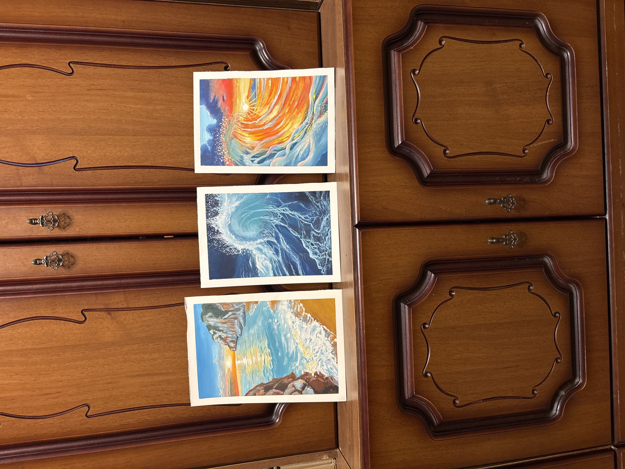 The image shows a close-up of a wooden dresser. Three watercolor paintings are placed on the dresser's surface. The top painting depicts an abstract swirl of orange, yellow, and blue. The middle painting shows a swirling blue wave. The bottom painting is a landscape featuring an ocean scene with rocks, a sunset, and waves crashing on the shore. The dresser itself has decorative trim around the drawers and ornate drawer pulls. No people are visible in the image. There is no indication of the location of the image being Izhevsk, Russia.