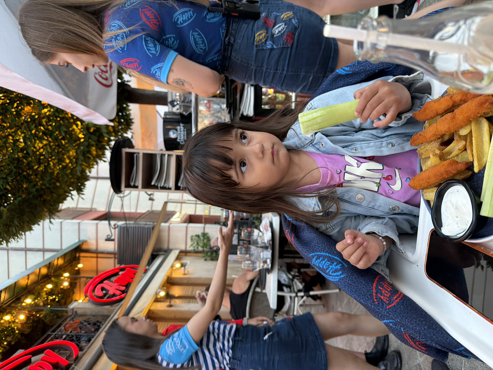 In a bustling outdoor restaurant setting in Burgas, Bulgaria, a young girl is seated at a high chair, holding a piece of celery. She is wearing a light blue denim jacket over a pink t-shirt, and a dark blue blanket with a repeating red and blue logo is draped over her. In front of her is a plate of food containing french fries and what appear to be chicken tenders, along with a small dish of white sauce. A smartphone is placed on the table in front of her. To her left, a woman wearing a blue and white striped t-shirt and a denim skirt is gesturing with her hand, possibly interacting with someone off-camera. To her right, another woman, wearing a blue t-shirt with repeating red and blue logos and a denim skirt, is standing and looking away. The background reveals the restaurant's facade with illuminated signs and overhead lights, as well as other patrons at tables. The overall atmosphere suggests a lively evening meal.