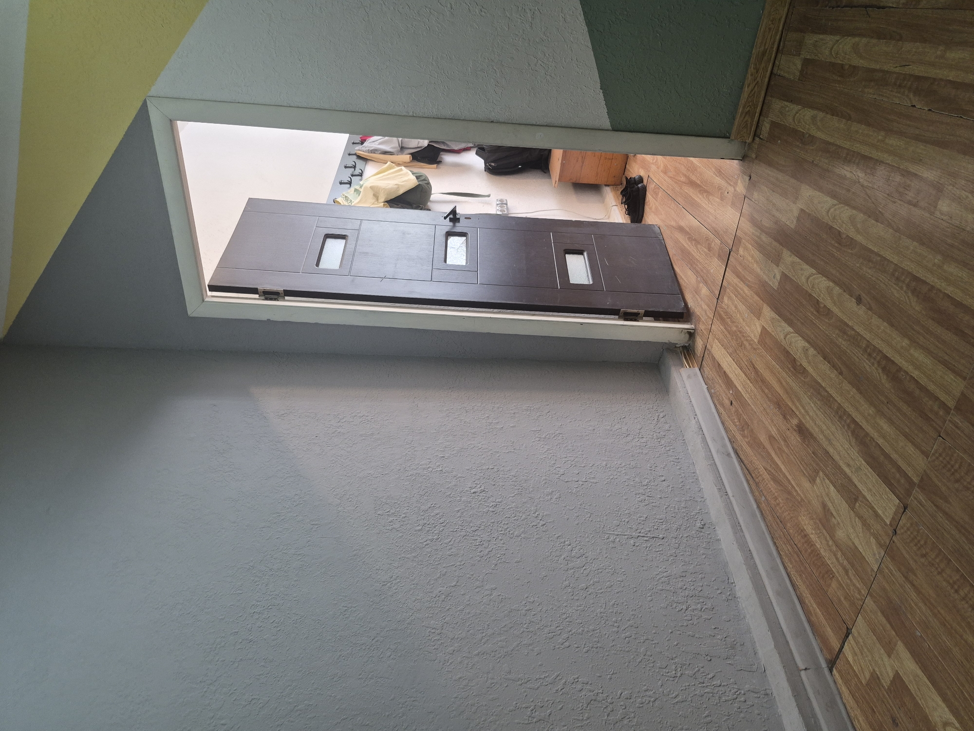 The image depicts a dimly lit interior hallway with a wooden laminate floor. To the left, a textured gray wall extends into the foreground, featuring a white baseboard. To the right, an open, dark brown door reveals a glimpse into an adjacent room. This room appears to be a small entryway or closet area, with a wall adorned with coat hooks from which several bags and articles of clothing are hanging. A small wooden bench sits against the back wall, and a pair of black shoes is placed on the floor in front of it. The walls of this adjacent room are painted in a light, neutral color. The hallway ceiling is partially visible, showing a diagonal color-blocked design with segments of yellow and green. This scene is likely from an apartment building hallway in Yerevan, Armenia, given the architectural style and typical interior finishes. There are no people visible in the image.