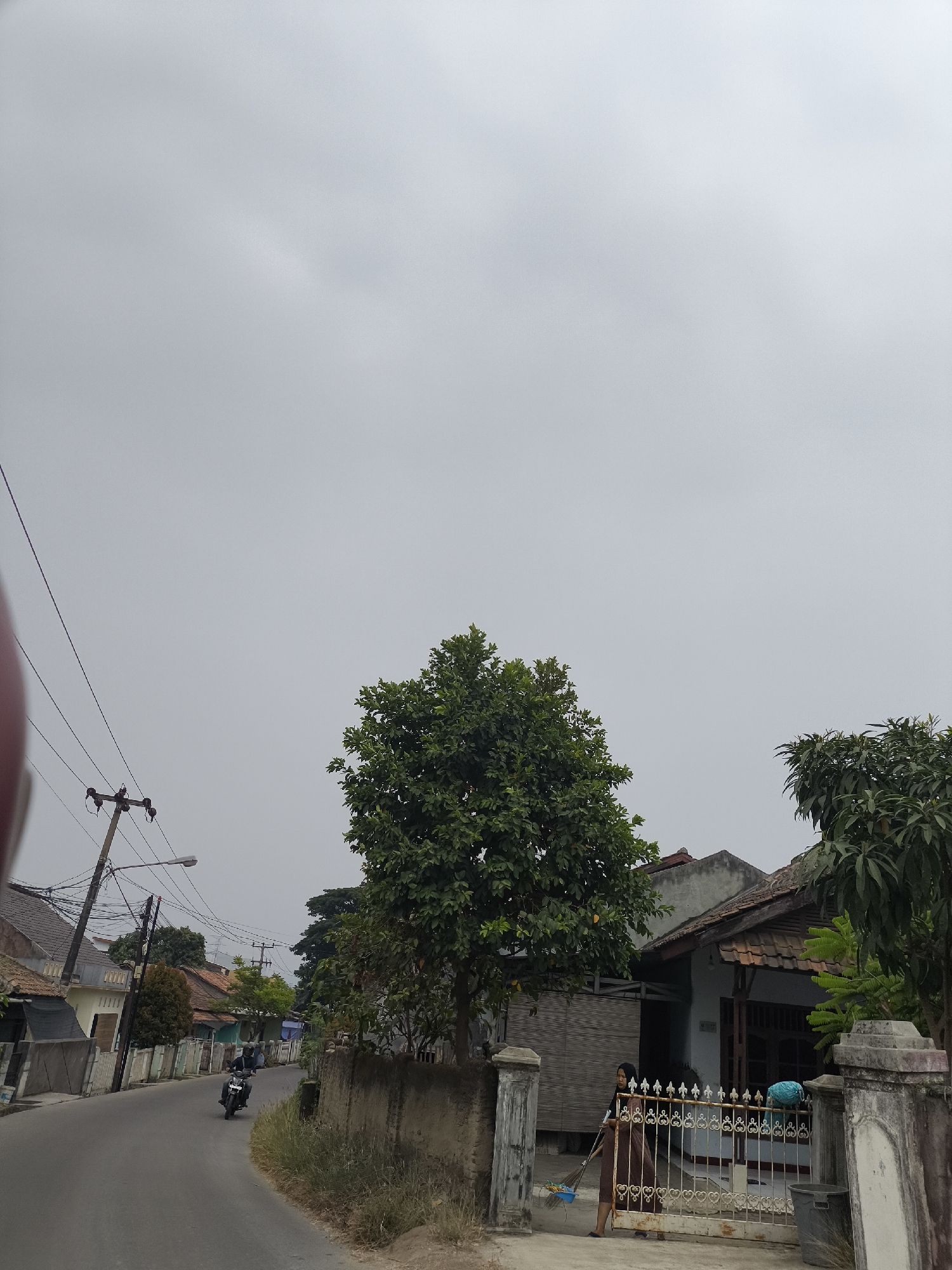 In Cilegon, Indonesia, a residential street is depicted under a cloudy sky. The road curves gently to the left, bordered by houses and vegetation. On the left side of the road, a motorcycle with a rider is moving away from the camera. Further up the street, there are several houses with tiled roofs and fences. Utility poles with electrical wires are visible along the side of the road. On the right side of the street, a house with a tiled roof is partially obscured by a large tree. In front of the house, a woman wearing a brown headscarf and dress is sweeping the ground with a broom and a blue dustpan. A white metal gate with decorative elements is in front of the house, and a trash bin is positioned next to the gate. The environment appears to be a typical suburban area with a mix of residential buildings and natural elements.