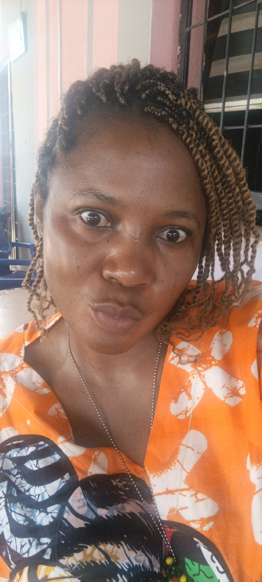 A medium shot shows a Black woman from the shoulders up, looking directly at the camera. Her skin is brown, and she has dark brown eyes and dark eyebrows. Her hair is styled in thin, brown braids that are pulled back from her face. She is wearing an orange shirt with a white, abstract pattern. A silver chain necklace with a small, colorful pendant is visible around her neck. Her lips are slightly pursed. The background is blurred but appears to be indoors, with vertical pink and white elements and a barred window visible on the right. The lighting suggests it is daytime.