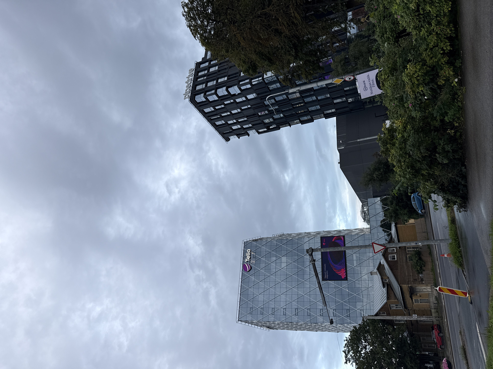 This image captures an overcast day in Tallinn, Estonia, showcasing a modern cityscape juxtaposed with older architecture. On the left, a tall, glass-covered building displays the "Telia" logo prominently at its peak. Below it, a large digital billboard shows a colorful abstract design and the Telia logo. To the right, a darker, more angular building features numerous windows and the "pipedrive" logo on its rooftop. In the foreground, a weathered, wooden house stands between the two modern structures, hinting at the city's layered history. A street with parked cars runs in front of the house, and trees and dense foliage partially obscure the view. The sky is filled with heavy, grey clouds, typical of Tallinn's climate. A traffic sign and a construction marker are visible near the street.