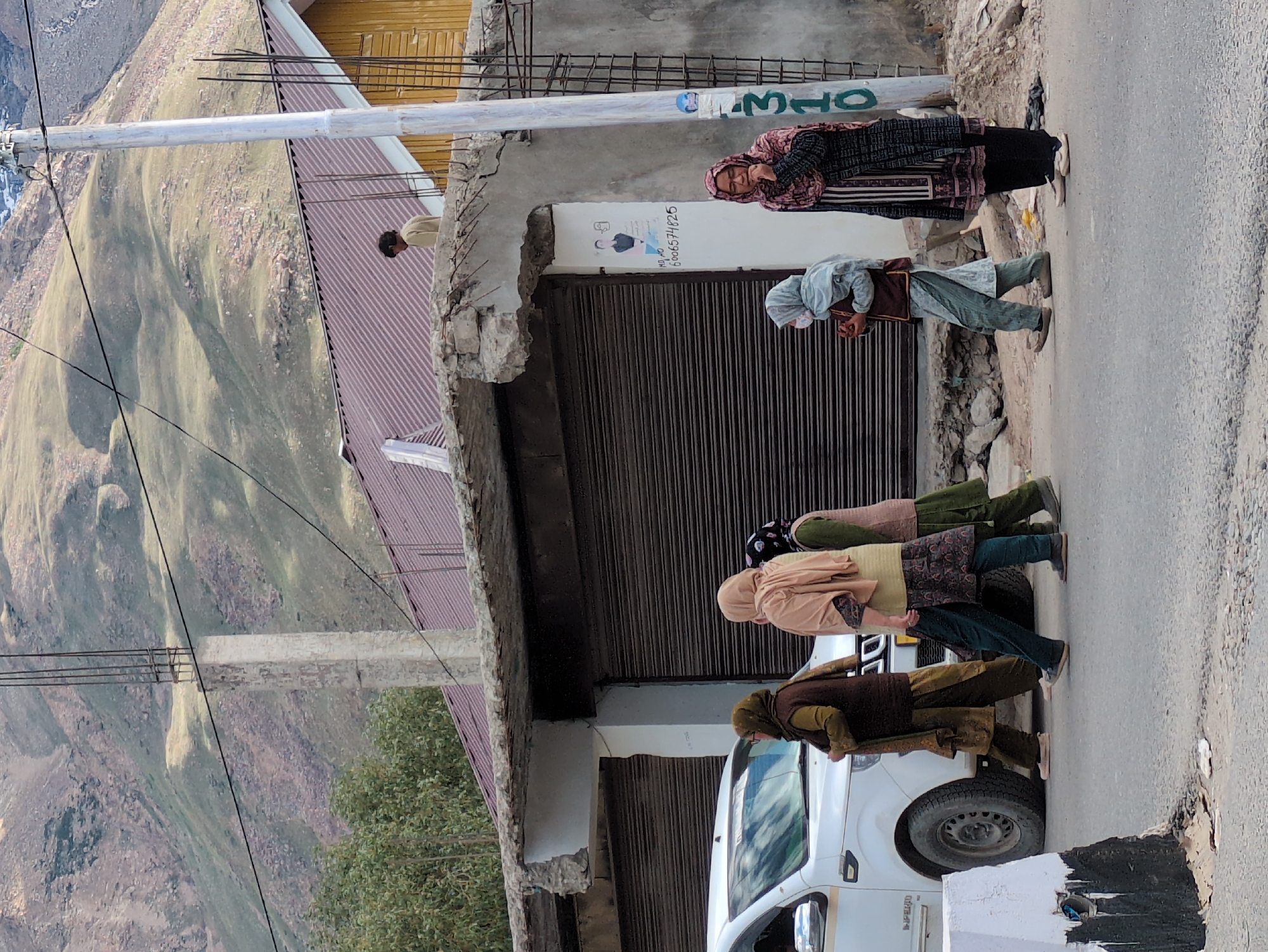 A group of five women are walking on a paved road in front of buildings. A white SUV is parked on the left side of the image. The buildings are made of concrete and have metal roll-up doors. In the background, there are mountains and trees. One woman is wearing a black and pink headscarf, a patterned long coat, and black pants. Another woman is wearing a light blue headscarf and a grey patterned outfit. A third woman is wearing a beige headscarf and a dark brown vest over a patterned tunic. A fourth woman is wearing a patterned headscarf and a patterned tunic with dark pants. The fifth woman is wearing a brown headscarf and a dark brown vest over a patterned tunic. A man is standing on top of a building in the background. The location is identified as Padum, India, based on visual cues and potentially external information.