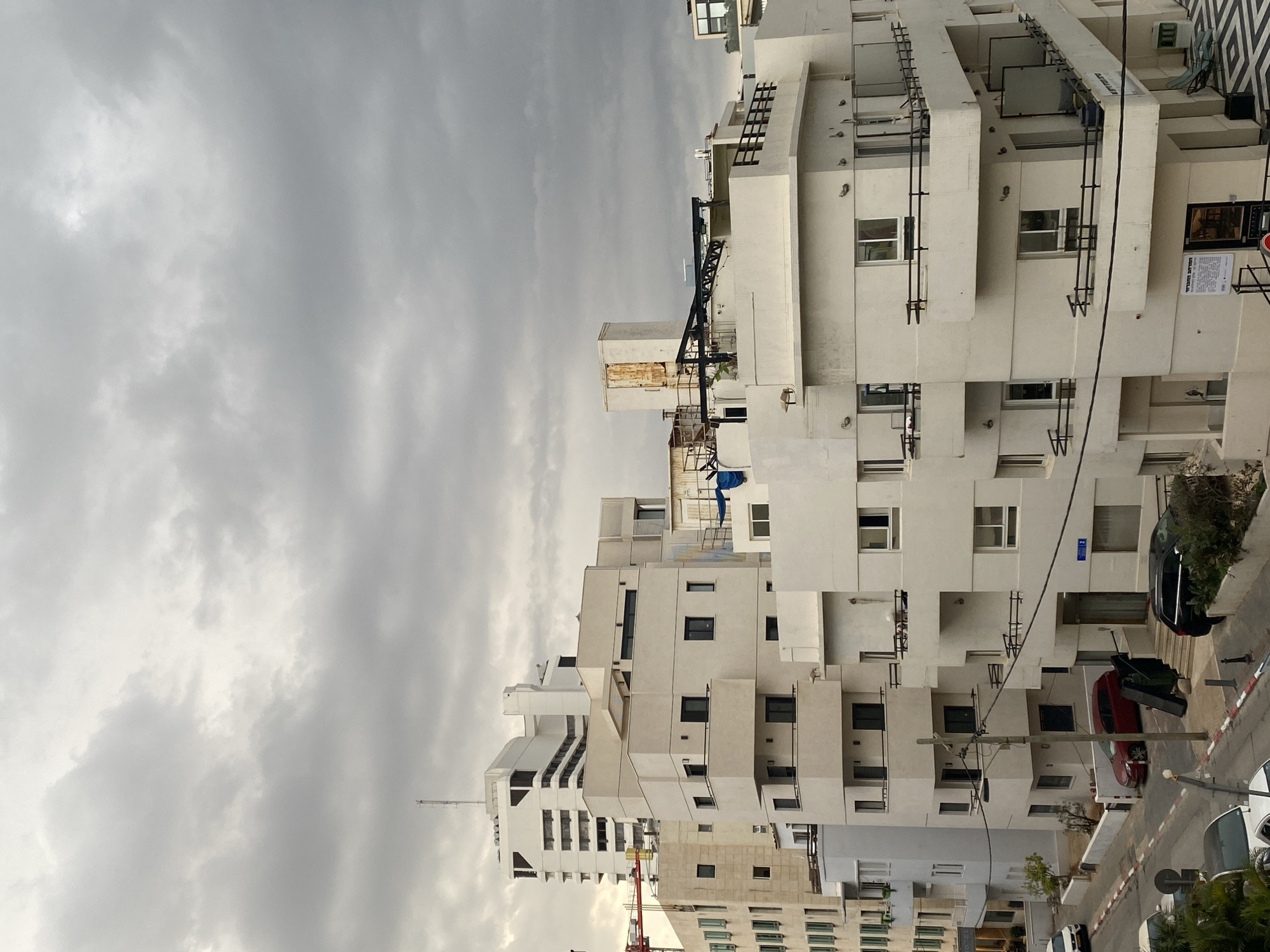 The image shows a cluster of white buildings against a cloudy sky. The architectural style features geometric shapes and stacked balconies. Cars line the street next to the building. The location is likely Tel-Aviv, Israel, given the architectural style characteristic of the city. There are no people visible in the shot. A construction crane is near one of the buildings.