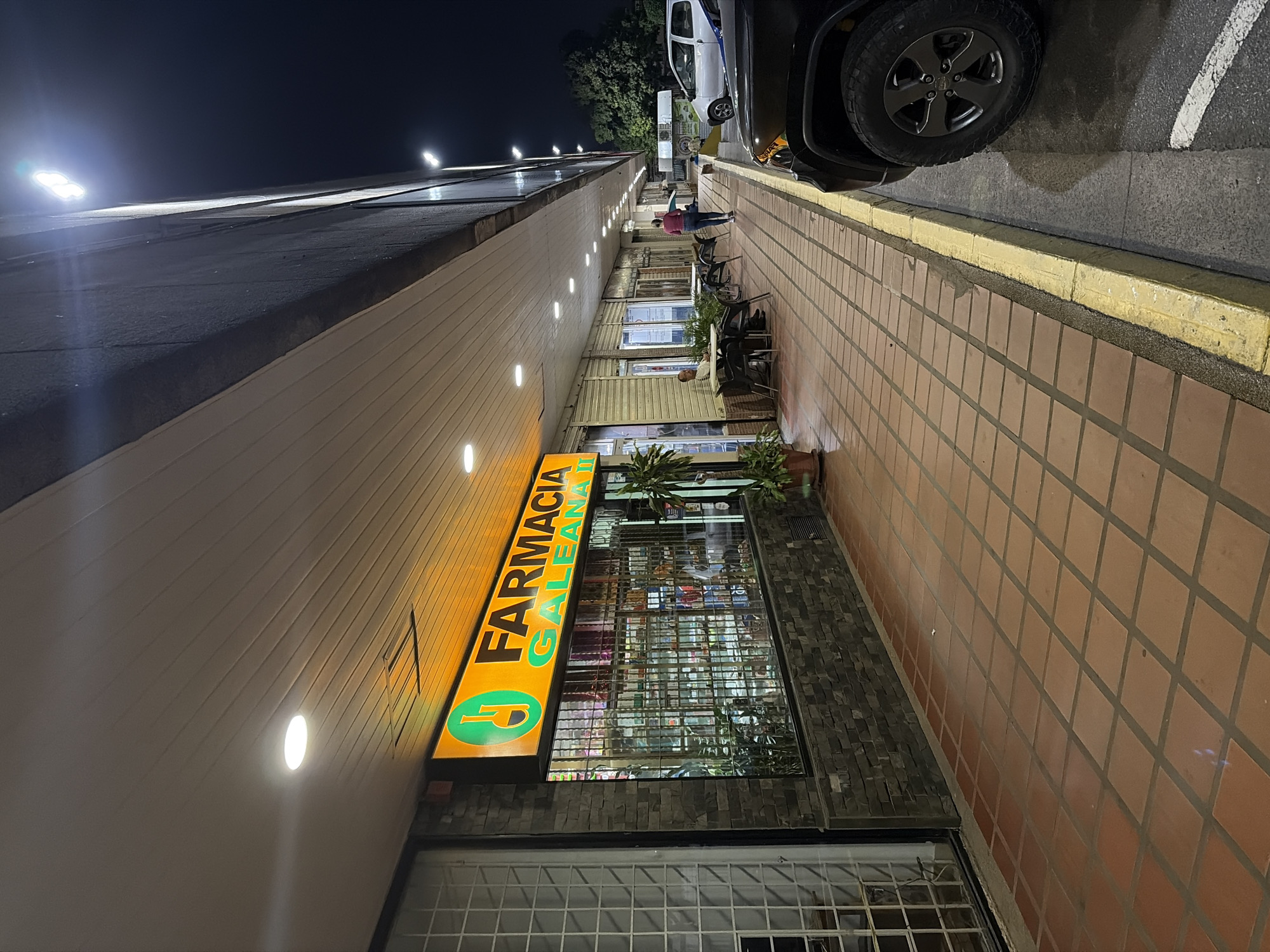 The image depicts a street scene at night in Guayana City, Venezuela. A "Farmacia Galeana" storefront is visible, with a bright yellow sign illuminating the facade. A tiled sidewalk stretches along the building, adjacent to a street where a black car and a white vehicle are parked. People are seated at tables outside what appears to be a restaurant or cafe along the sidewalk. The environment is dimly lit with street lights.