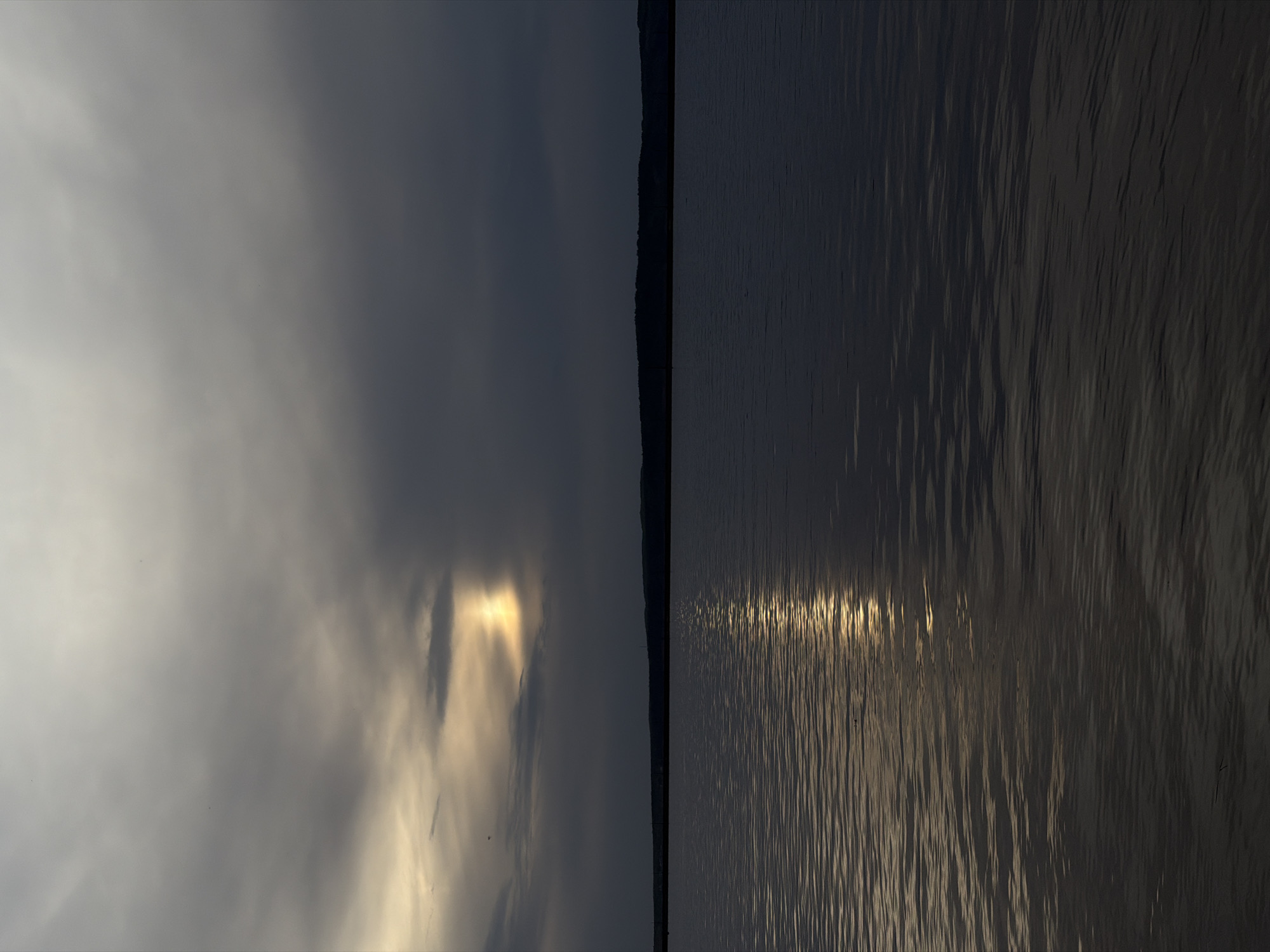 The image depicts a waterscape, divided vertically into two distinct halves. The left side features a cloudy sky with a break in the clouds allowing sunlight to peek through. The right side displays a body of water, possibly a river or lake, with sunlight reflecting off its surface, creating a rippled, shimmering effect. A dark, thin strip of land is visible between the two halves. This scene is likely located in or near the city of Chaikovsky, Russia, given the prompt's specification, but the image itself provides no explicit evidence to confirm this location.