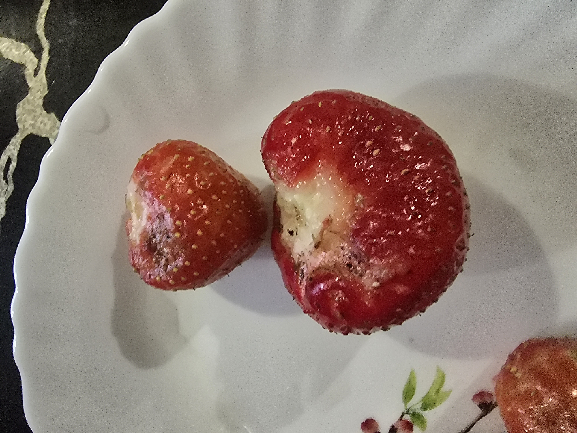 Two partially eaten strawberries are arranged on a white, scalloped plate. One strawberry, to the left, appears to be intact with visible seeds on its surface. The other, larger strawberry, to the right, is significantly damaged, with a large portion of its flesh missing, revealing the white interior. Both strawberries are a vibrant red color. A portion of another red fruit, possibly a third strawberry, is visible in the bottom right corner of the frame. The plate rests on a dark, textured surface that has a swirling pattern, suggesting a table or countertop. Faint shadows are cast on the plate, indicating the presence of light from above and to the left. The overall impression is of a simple still life with a focus on the strawberries and their condition. There is no visible text in the image. The context of Chebarkul, Russia, does not provide specific visual cues within the image itself.