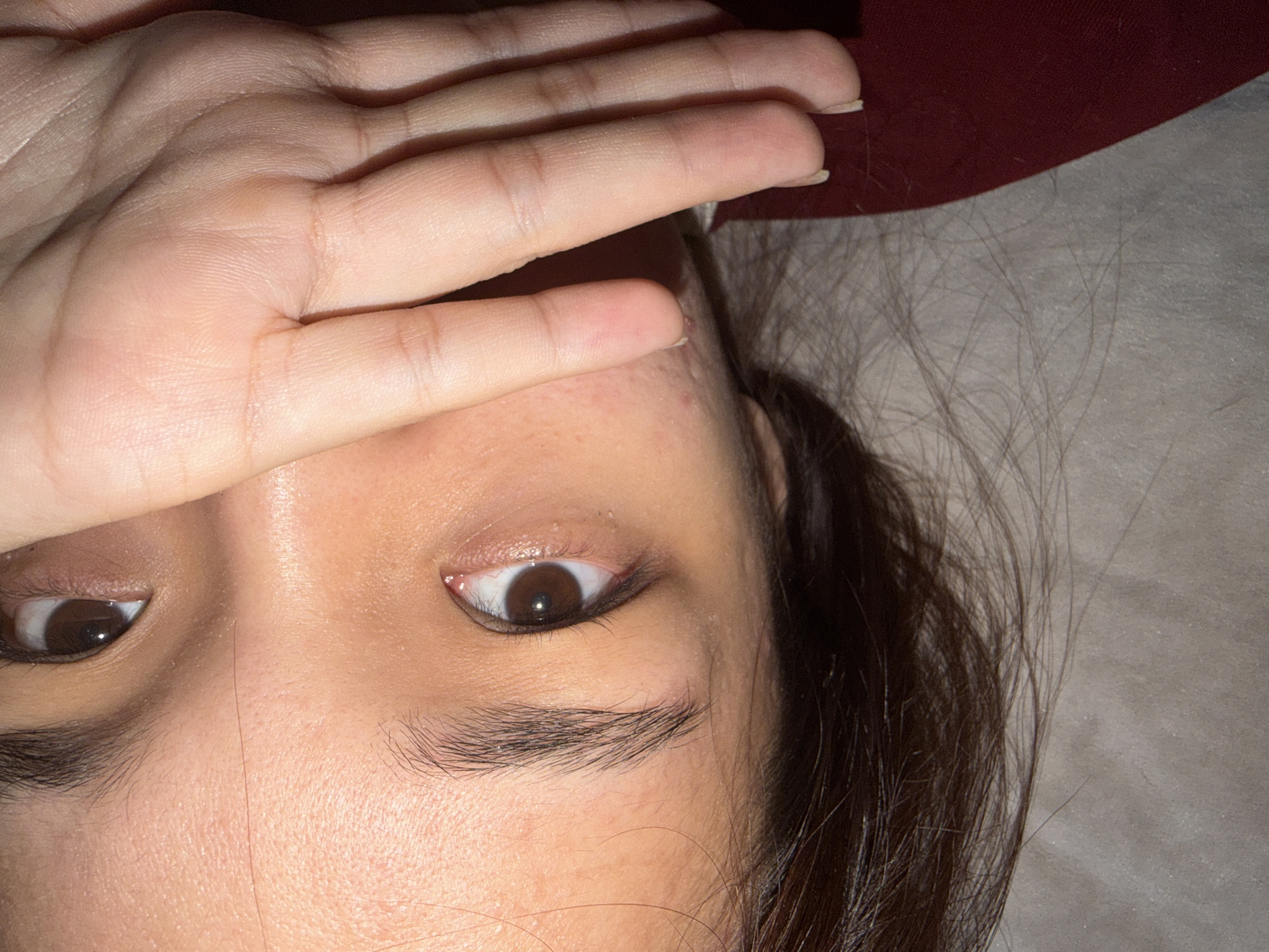 A close-up, over-the-shoulder shot focuses on the lower half of a person's face, including one eye and part of their forehead. Their hand is raised, covering their mouth and nose. The person's skin appears to have some texture and minor blemishes. The background is out of focus, showing a muted, textured surface. A portion of a dark red garment is visible at the bottom left of the frame.