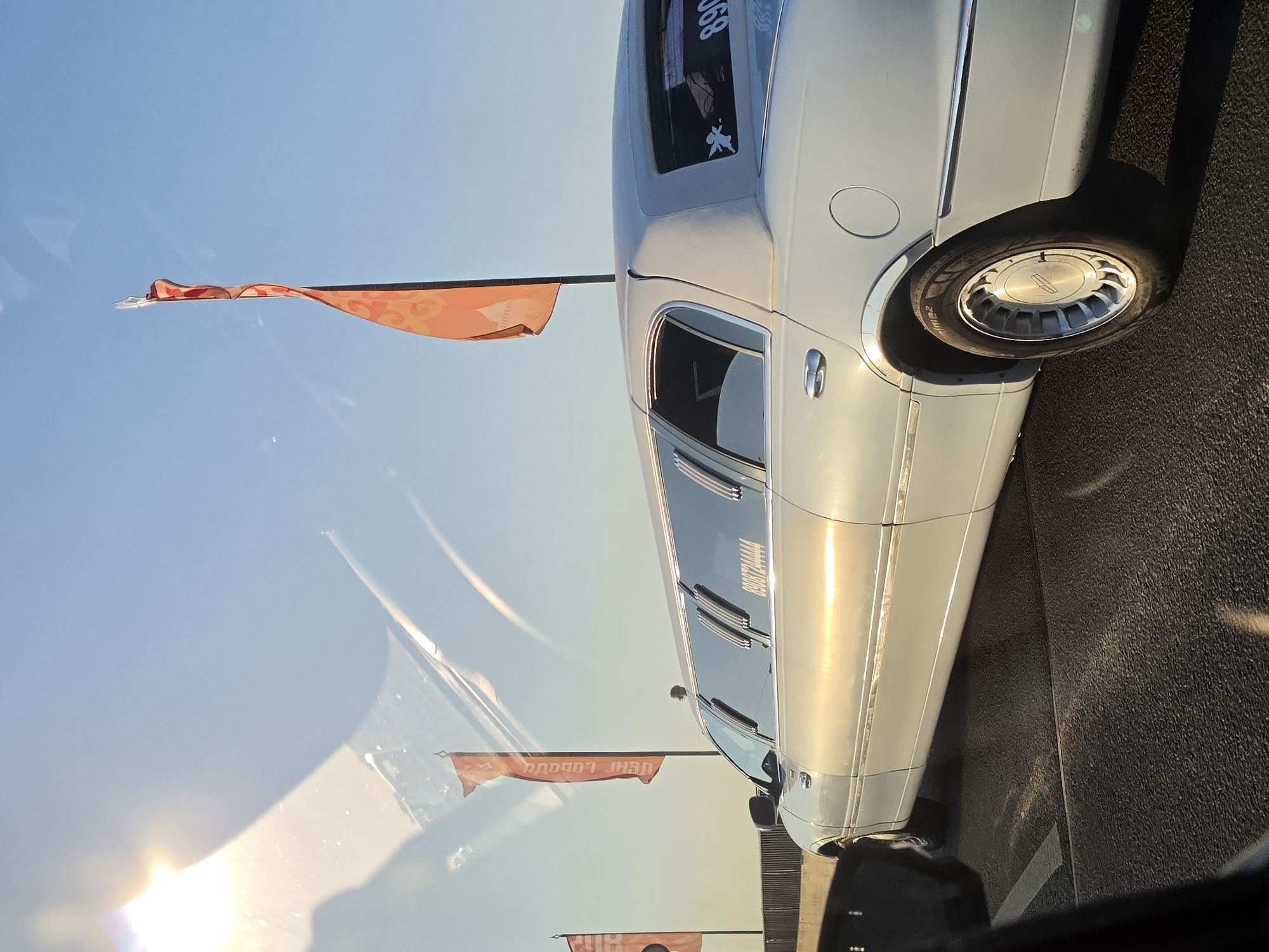 An extended white limousine drives along an elevated road under a clear blue sky, with the sun's rays creating glare on the vehicle and the road surface. Orange banners with stylized patterns are visible on poles along the roadside, and a faint reflection of the sun can be seen on the limousine's windows. The limousine has several tinted windows along its length, and a phone number "89037244444" is printed in white on the rear passenger window, along with a small white sticker of an angel. The road appears to be asphalt with lane markings. The background suggests an urban or semi-urban environment, with a bridge-like structure on the left and a railing along the side of the elevated road. The scene is likely captured from inside another vehicle. Given the context of flags and specific phone number, it's possible this is part of a promotional event or transportation service in Nizhny Novgorod, Russia.