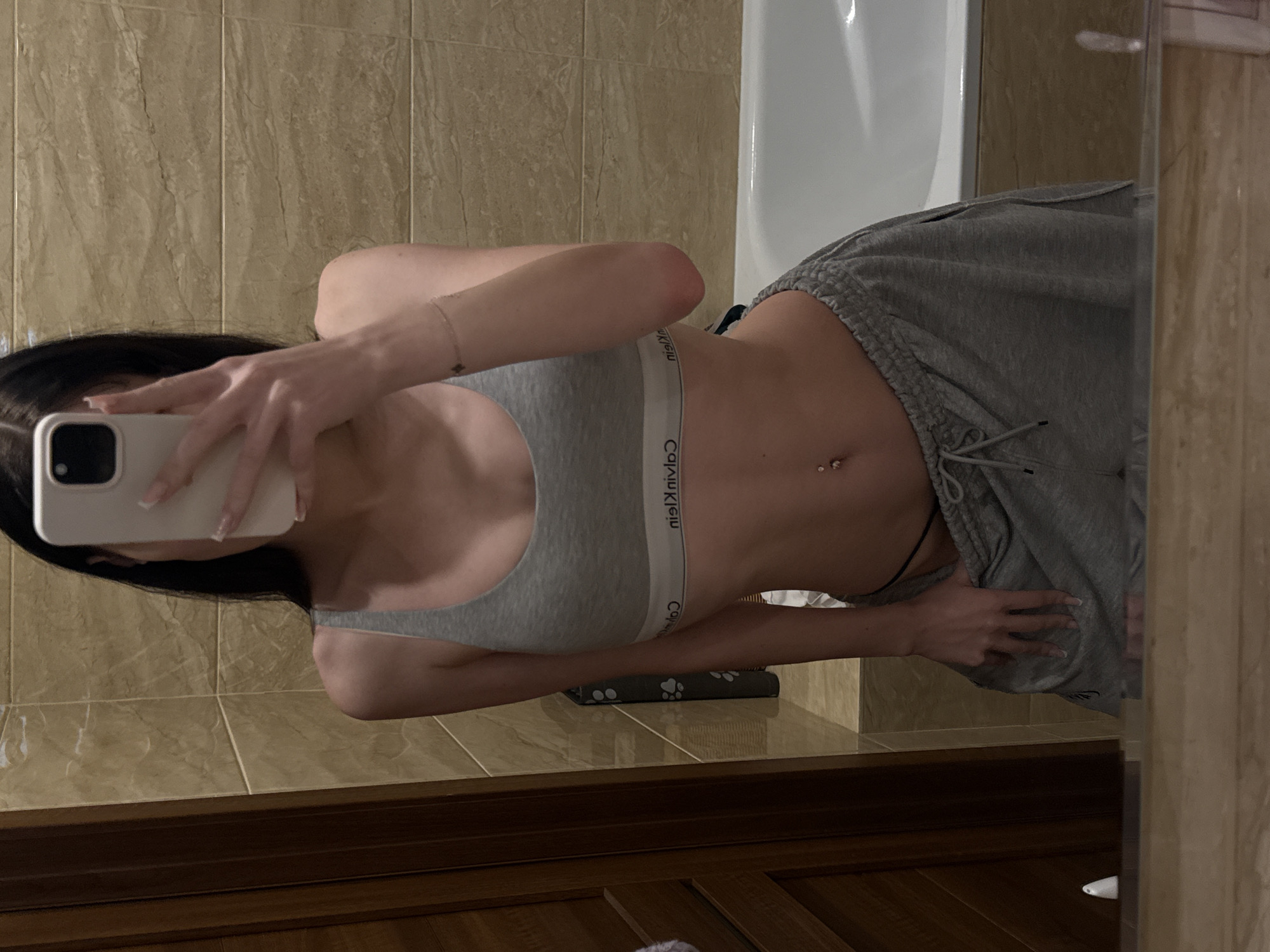 The image is a mirror selfie taken by a person in a bathroom. The person is wearing a grey Calvin Klein sports bra and grey sweatpants. They have a belly button piercing. The background shows beige tiled walls and a white bathtub. There is a wooden cabinet below the mirror. The lighting suggests it is likely daytime or a well-lit interior. The overall mood is casual and relaxed. The text "Calvin Klein" is visible on the waistband of the sports bra.