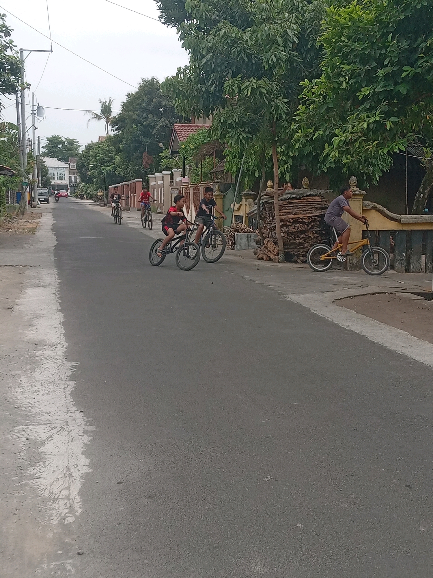 In Maguwoharjo, Indonesia, a street scene unfolds with several children riding bicycles. In the foreground, a boy wearing a red shirt and dark shorts rides a black BMX-style bicycle. To his right, another boy in a black t-shirt and dark shorts is also on a bicycle. Further down the road, two more children are visible on bicycles, one in a red shirt and the other in a black shirt. On the right side of the street, a young man in a gray t-shirt and shorts cycles a yellow bicycle. The road surface is asphalt, with a lighter, wet-looking patch on the left side. Buildings line the right side of the street, featuring beige walls and red-tiled roofs, interspersed with lush green trees and foliage. A pile of stacked lumber is visible near a fence on the right. In the background, more trees and buildings are present, with utility poles and wires overhead. The sky is overcast.