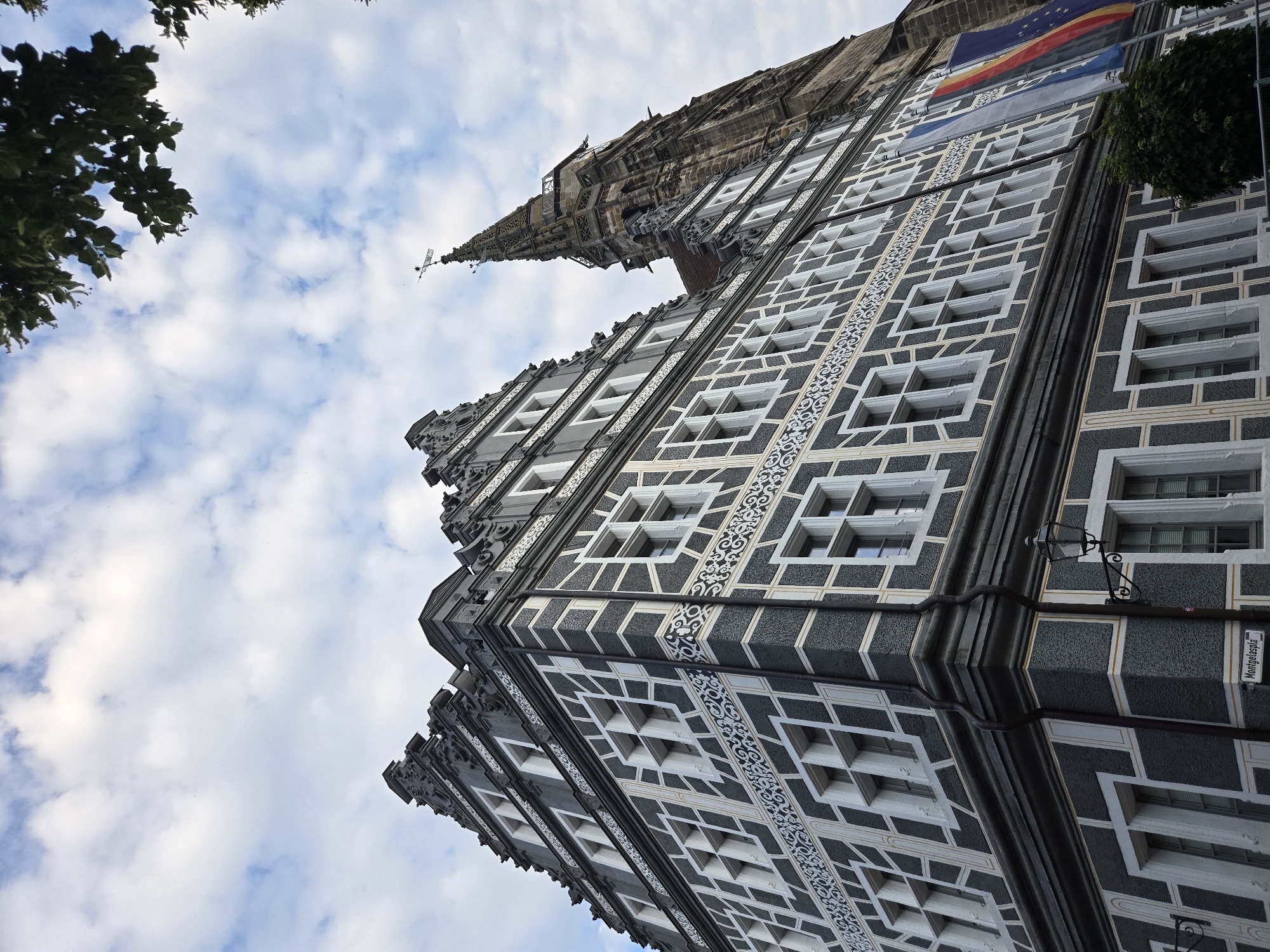 This is a low-angle, eye-level shot of the Ansbach Town Hall (Rathaus Ansbach) in Ansbach, Germany, taken on a partly cloudy day. The focus is on the ornate facade of the historic building, characterized by its dark grey plaster with a distinctive white geometric pattern, resembling half-timbering. Multiple rows of windows with white frames punctuate the facade. To the right, a tall, Gothic-style tower, likely part of a church, rises against the sky. German and European Union flags hang on the side of the building near the tower. Green foliage from a tree is visible in the upper right corner of the image, and a street lamp is mounted on the building's facade. A street sign is partially visible at the bottom left, reading "Montgelaspla".