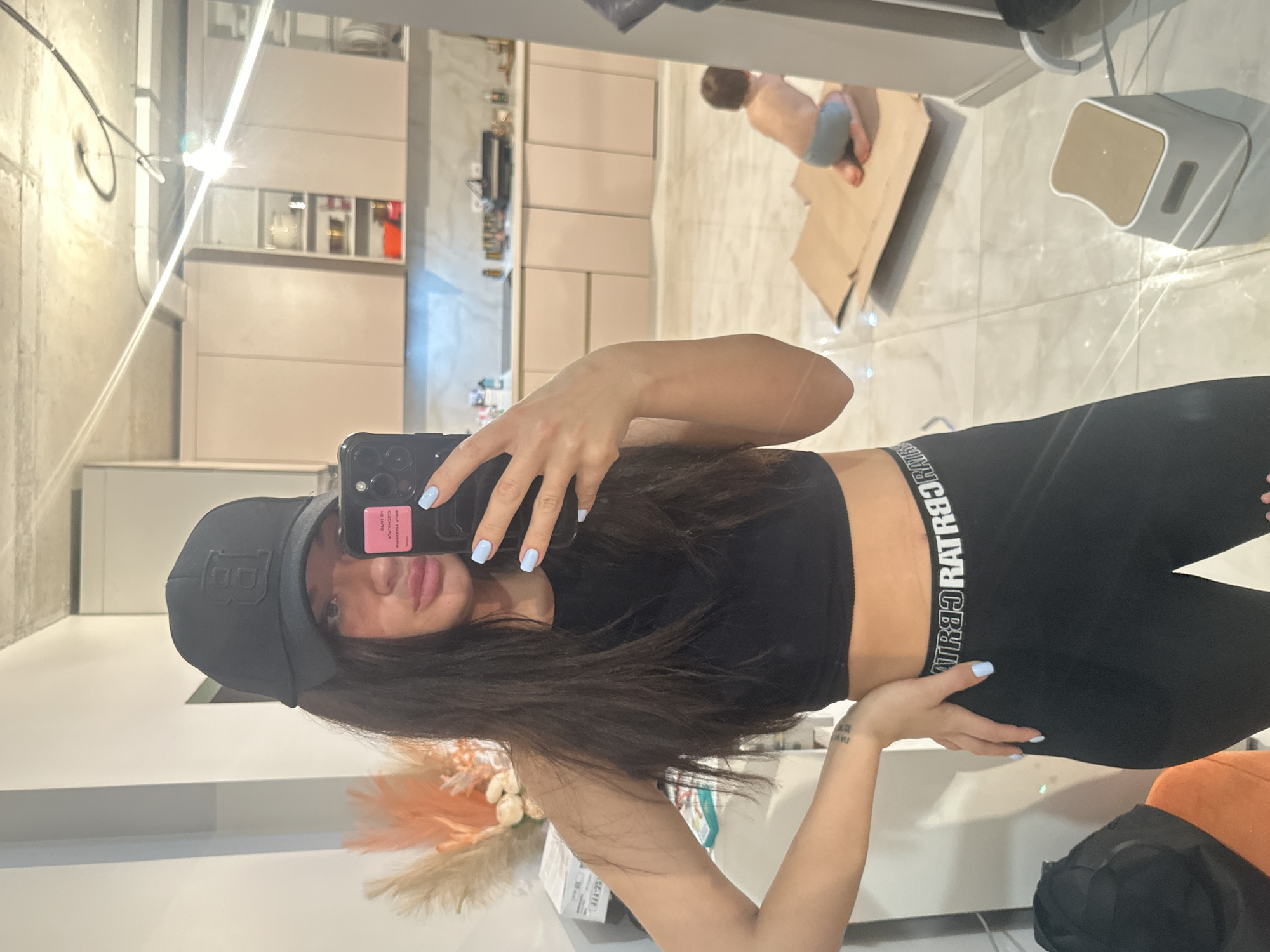 A woman in a black crop top and black leggings with white lettering on the waistband is taking a selfie in front of a mirror. She is wearing a black baseball cap with a white "B" logo. Her dark hair partially covers her face, and she has light blue nail polish. The reflection shows a kitchen with light-colored cabinets and marble countertops. A child is visible in the background, shirtless and wearing blue shorts, playing with a cardboard box. The floor is a light marble pattern. In the foreground is a floral arrangement in a vase. The location is in an apartment. There is no information in the image that indicates the location is in Khimki, Russia.