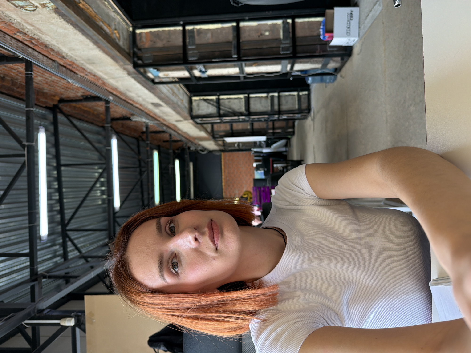 An indoor, eye-level shot in an industrial-style office or co-working space located in Chaikovsky, Russia. A woman, appearing to be in her mid-20s to early 30s, is seated at a white desk in the foreground, looking directly at the camera with a neutral expression. She has light red, shoulder-length hair styled in a straight bob and is wearing a white ribbed short-sleeved t-shirt and a thin black necklace. Her right forearm rests on the desk surface.
The background reveals a large open-plan area with an exposed ceiling featuring dark metal structural beams and corrugated metal panels, illuminated by multiple long, horizontal fluorescent tube lights. Exposed red brick walls are visible along the upper right side. A series of dark-framed partitions or room entrances with light-colored panels line the right side of the space, extending into the distance. The floor is light grey, consistent with concrete. In the deeper background, a blue water cooler jug and white cardboard boxes, one marked "COPY," are discernible. The overall environment suggests a functional workspace.