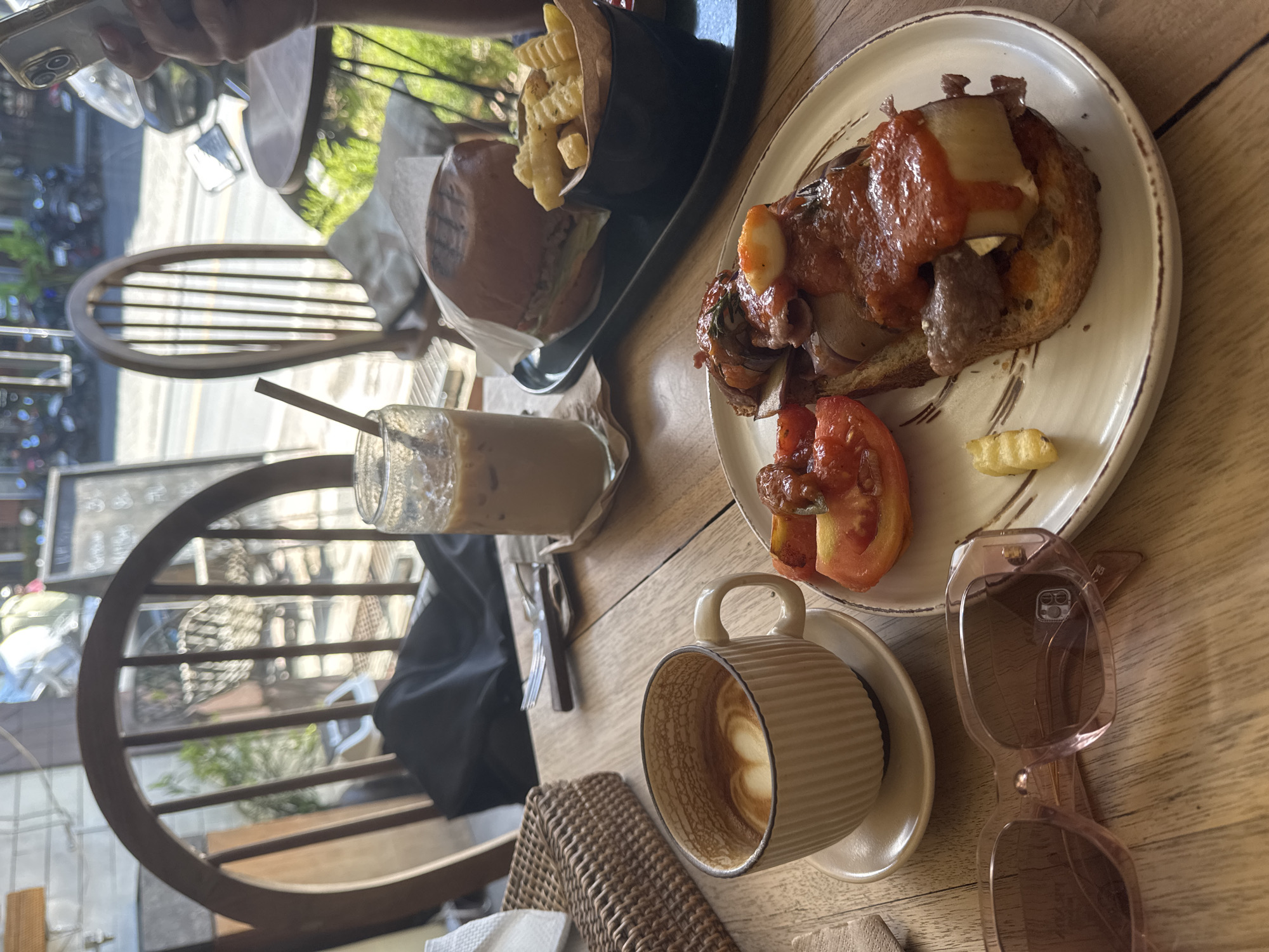 A slightly high-angle, full shot depicts a cafe table with a variety of food and drink items. The table has a natural wood grain finish. Dominating the upper right of the frame is a white ceramic plate holding a slice of toast topped with tomato sauce, mushrooms, and a slice of tomato on the side. Beside the plate is a pair of pink-framed sunglasses. In the center of the frame, a light brown ceramic mug filled with latte art rests on a saucer. Next to it is a glass of iced coffee with a straw. To the left, a black container holds french fries, next to a wrapped burger. A wicker-backed chair is positioned on the lower left of the image, with an outdoor cityscape visible through the back of the chair and the cafe window. An open container holding napkins is visible on the lower left. A partial view of another table and chair can be seen in the upper left. The location is a cafe in an urban area.