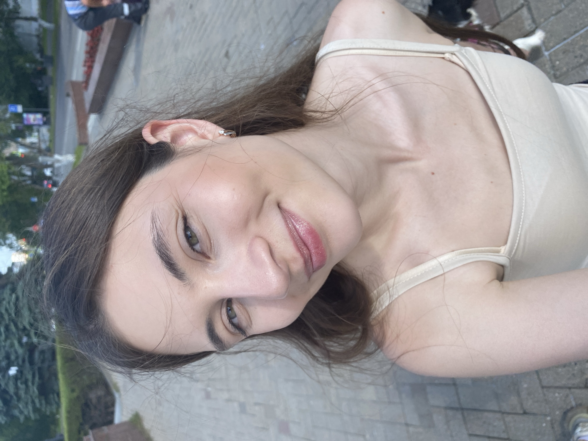 The media file is a portrait of a young woman, taken outdoors in Izhevsk, Russia. The woman has long, dark brown hair and is wearing a light-colored tank top. She is looking towards the camera with a slight smile. Her makeup includes lipstick and eyeliner, and she has a small earring in her left ear. The background shows a paved area with some greenery and buildings in the distance, suggesting an urban environment. The lighting suggests it is daytime, possibly late afternoon or early evening, with soft, diffused light. The orientation of the image is portrait, with the subject tilted.