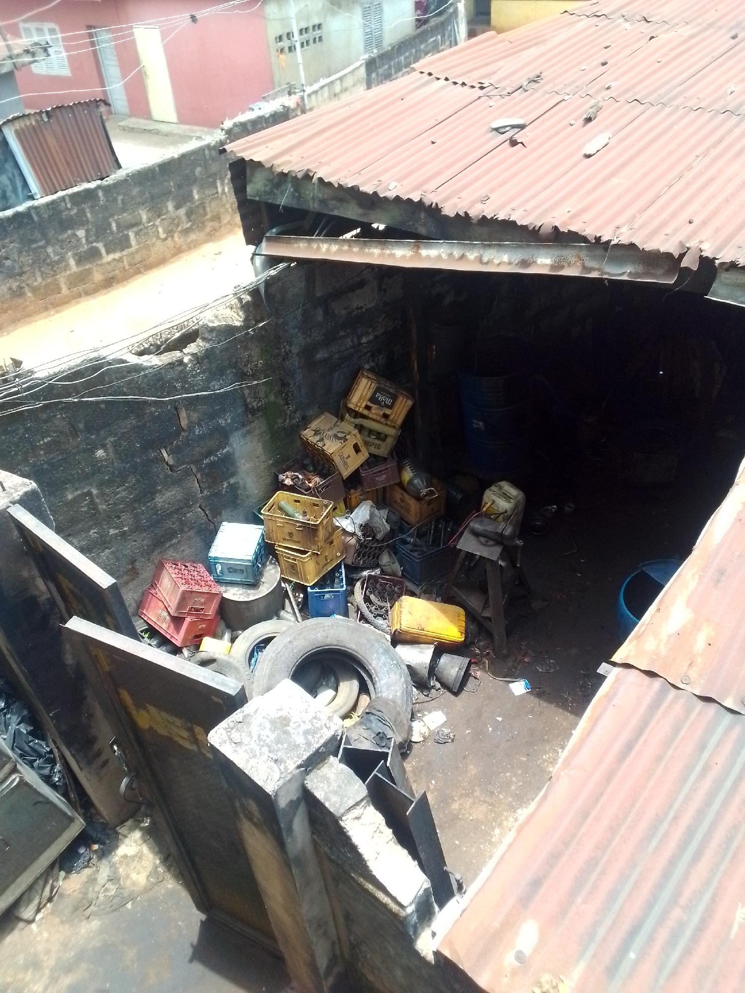 The image depicts a cluttered, outdoor workspace in an urban setting, likely in Unknown, Unknown. The area is enclosed by weathered concrete walls and covered by a corrugated metal roof. Various objects are scattered throughout the space, including several stacked plastic crates of different colors (red, blue, yellow), used tires, metal containers, barrels, and a wooden workbench. The overall impression is one of disarray and a lack of organization. The lighting is dim inside the workspace, making it difficult to discern details. In the background, beyond the walls, there are other buildings, suggesting a densely populated neighborhood.
