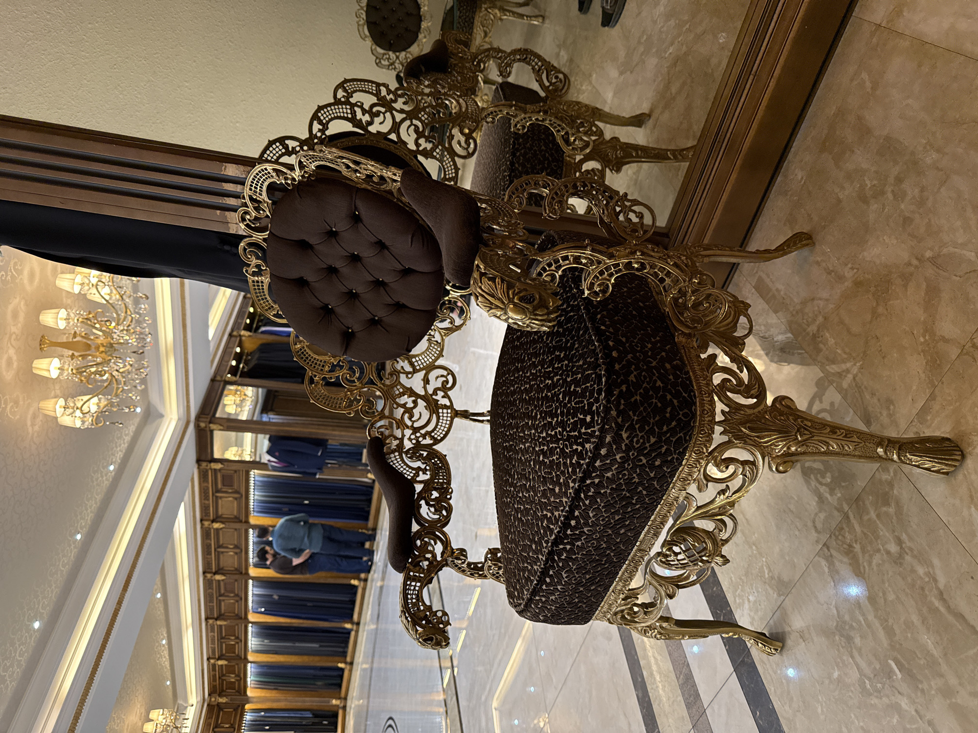 This image depicts the interior of a high-end tailor shop, likely located in Baku, Azerbaijan. The foreground is dominated by an ornate, gilded armchair with dark brown tufted upholstery on the back and a leopard-print patterned seat. The chair's frame is elaborately carved with swirling motifs and decorative elements. In the background, two men are seen standing in front of a display of fabric rolls, suggesting they are either customers or staff engaged in selecting materials. The shop appears luxurious, with a polished marble floor, intricate ceiling details, and chandeliers, indicating a sophisticated ambiance. A large mirror reflects the scene, adding depth and showcasing the shop's refined interior. The overall impression is one of opulence and bespoke tailoring services.