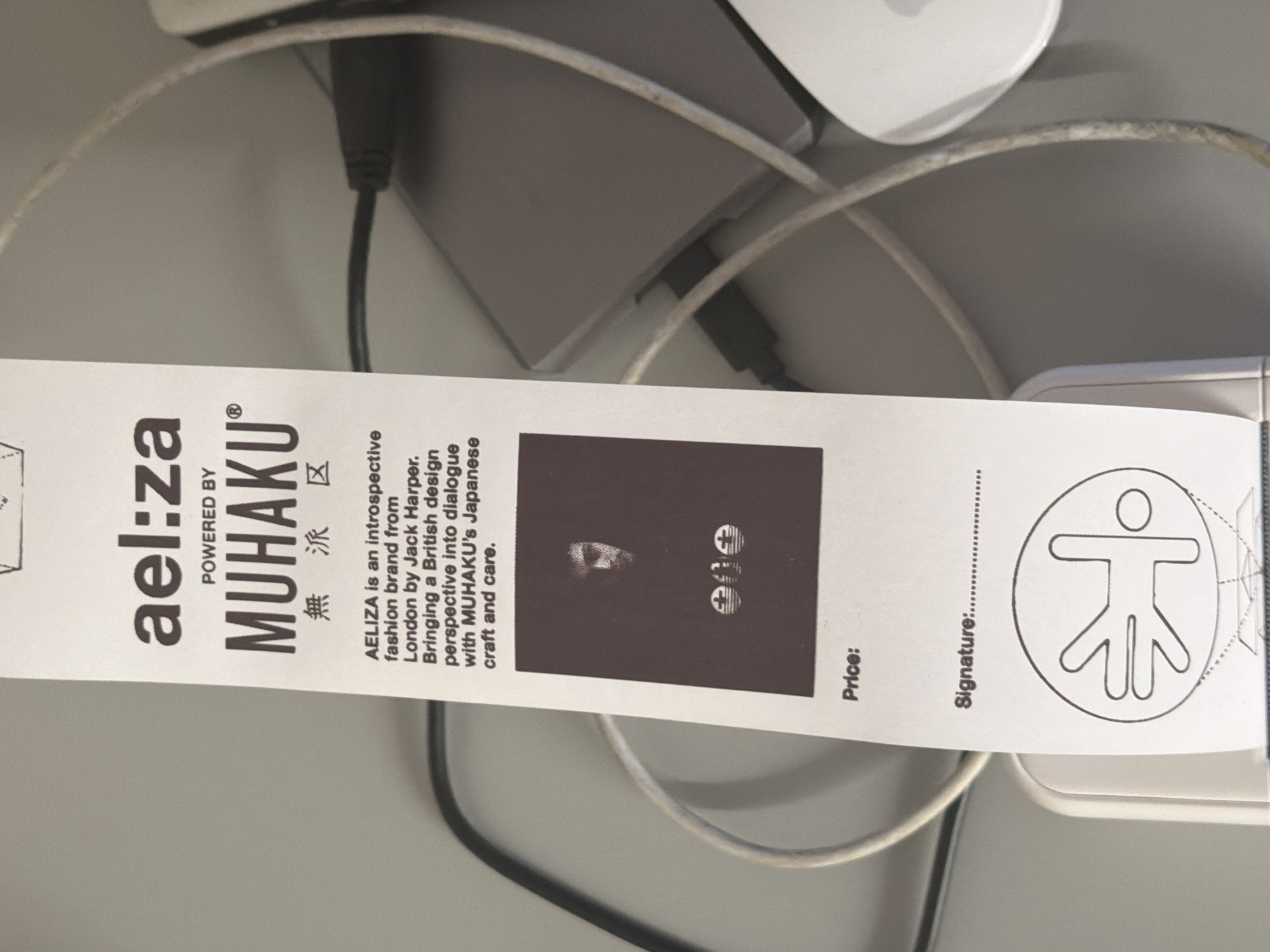 The image shows a close-up of a white tag with black text and graphics. The tag is printed with the brand names "ael:za" and "MUHAKU," and includes a brief description of AELIZA as a fashion brand from London by Jack Harper, combining British design with MUHAKU's Japanese craft. There is also a black and white picture. The tag also has sections labeled "Price:" and "Signature:," and a graphic of a stick figure within a circle. The tag is placed on a surface with various cables, a laptop, and a white mouse visible in the background. There is no indication of where the image was taken.
