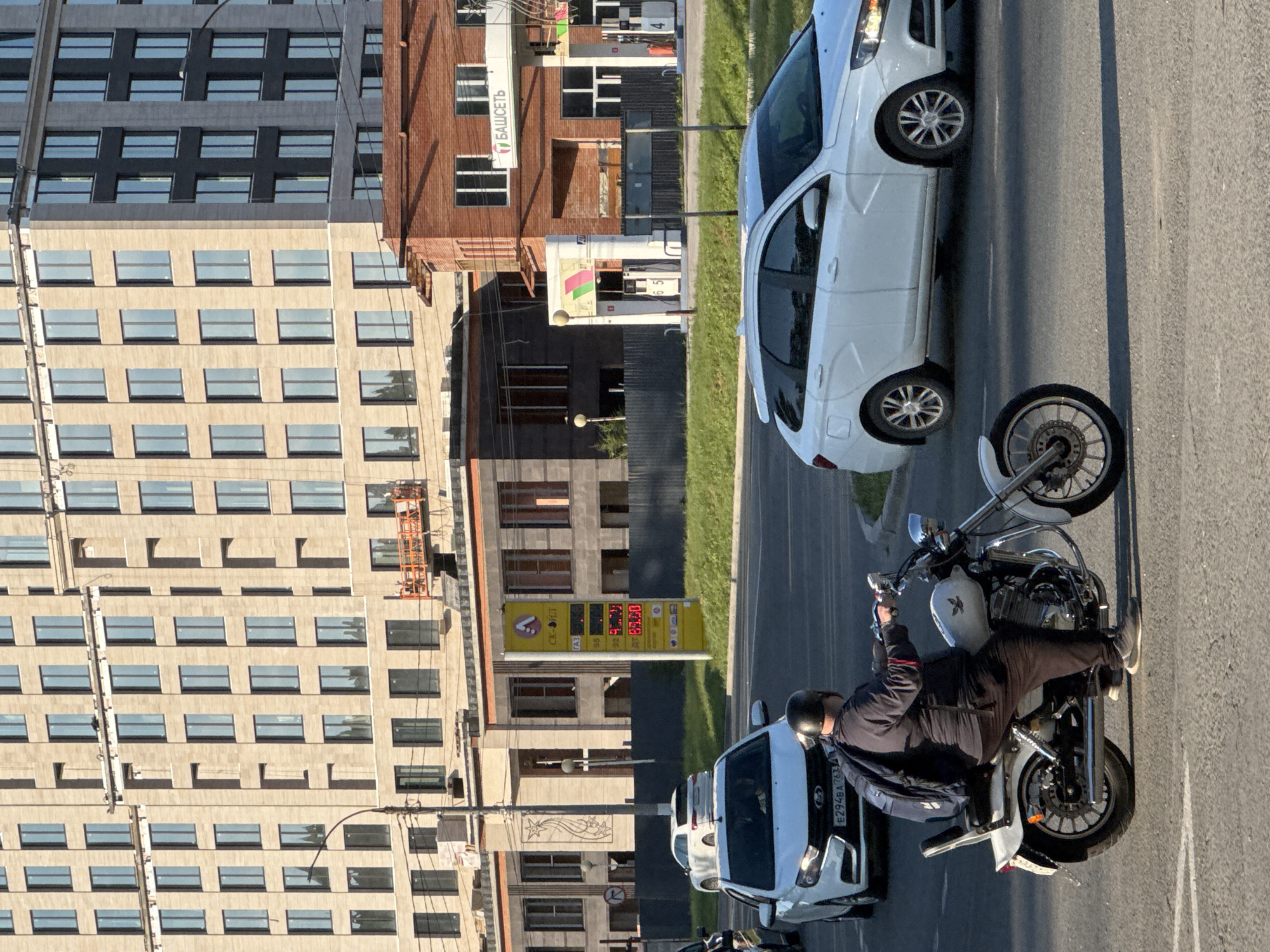 The image shows a street scene with a tall, modern building on the left and a gas station on the right. In the foreground, a person is riding a motorcycle. The motorcycle is silver and black, with chrome accents. The rider is wearing a black jacket, helmet, and dark pants. The motorcycle is positioned on the asphalt road, which has white lane markings.
Next to the motorcycle, a white car is parked. This car is a modern sedan, and its license plate is partially visible. Behind the car and motorcycle, there is a gas station with multiple pumps and signage. A prominent yellow sign displays fuel prices, including numbers like "91.99", "92", "95", and "98.30". The gas station building appears to be constructed with brick and glass.
Further in the background, the large building on the left features many windows arranged in a grid pattern. Some windows are dark, while others reflect the sky and surroundings. The building's facade is a mix of light-colored stone and darker sections. The overall scene suggests an urban or suburban environment.
The lighting indicates it is daytime, likely late afternoon or early evening due to the long shadows cast by the building and the warm, golden hue of the light. The sky is not clearly visible, but the sunlight suggests fair weather. The activity depicted is the normal flow of traffic and activity at a gas station. No specific emotions are discernible.
Visible text includes pricing information on the gas station sign and a brand name that appears to be "БИЗОТ". There are also some Russian characters on the license plate of the white car, starting with "E294BA".