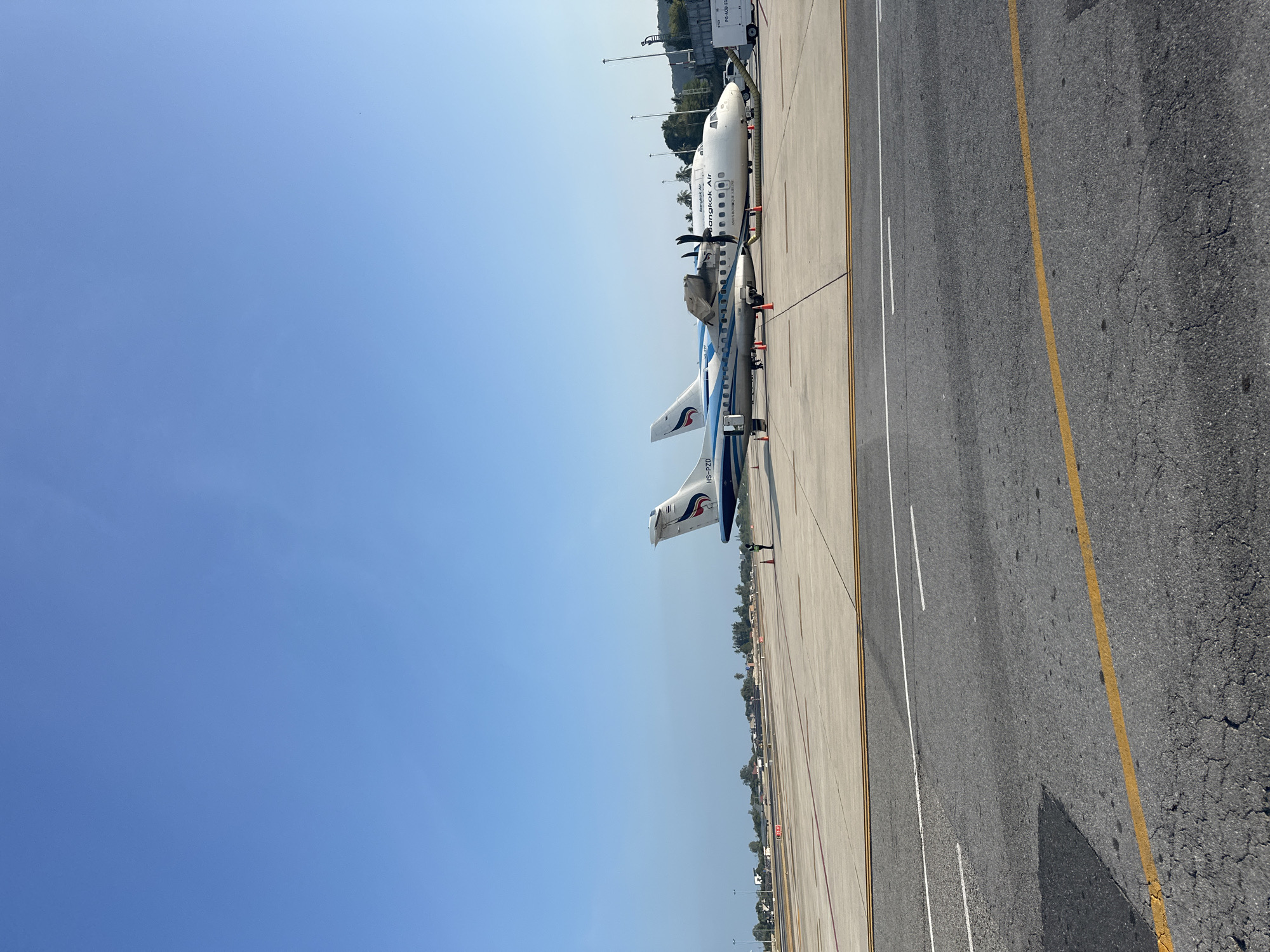 The image shows two Bangkok Airways airplanes parked on a tarmac. The location appears to be an airport in บ้านบางรักษ์, Thailand, based on the airline branding. The sky is clear and blue. The airplanes are positioned parallel to each other. The tarmac has painted lines. A section of an asphalt road is visible on the right.