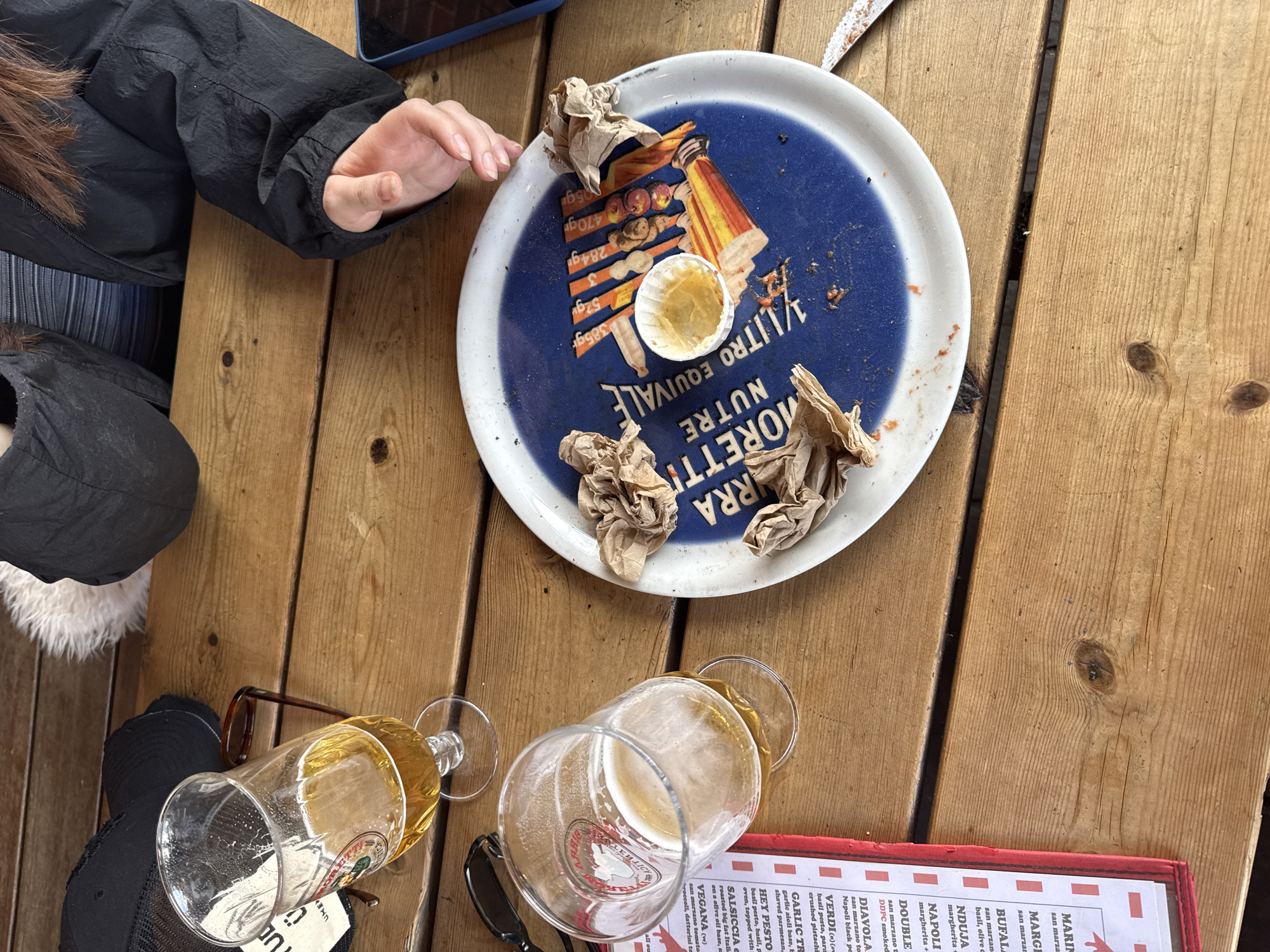 The image shows a wooden picnic table. A person wearing a black jacket is visible in the upper left. On the table is a white and blue tray with paper napkins and a small container. Two empty beer glasses are on the table, along with a menu card and a pair of sunglasses. A shoe and a hat are also visible. The OCR text suggests the menu is for a pizza restaurant and includes a list of pizzas such as Margherita, Bufala, Nduja, Napoli, and Diavola. The text also indicates the trays are for Moretti beer. The image provides no data to show where this image may have been taken in North Hertfordshire.
