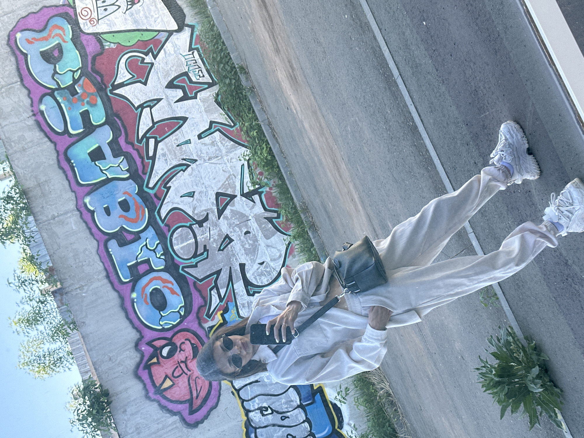 A full shot shows a person standing on an elevated concrete ramp next to a graffiti-covered wall. The person is facing forward and taking a selfie with their phone. They are wearing a cream-colored sweatsuit, white sneakers, and black sunglasses. A black purse hangs from their shoulder. The wall to their left is covered in graffiti in a variety of colors including purple, blue, red, and white. Some green foliage grows out from behind the wall. To the right of the person is a section of concrete and some more foliage. The photograph's orientation makes the ramp appear to be at an angle. Based on the graffiti and general environment, this may be located in the city of Izhevsk, Russia.