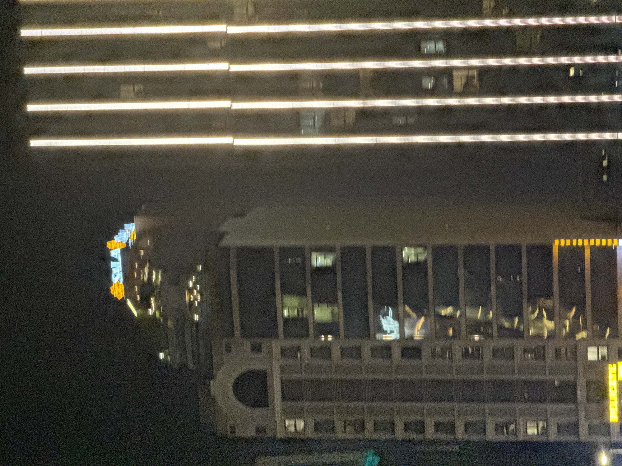 The image shows a nighttime view of buildings in Đà Nẵng, Vietnam. A tall structure with horizontal illuminated lines spans the upper portion of the frame. Below it is a lower, wide building with a grid of windows, some showing interior lights. The lower building has a distinctive archway feature. To the left, an additional building with illuminated signage and architectural details is visible against a dark sky. The image is slightly tilted.