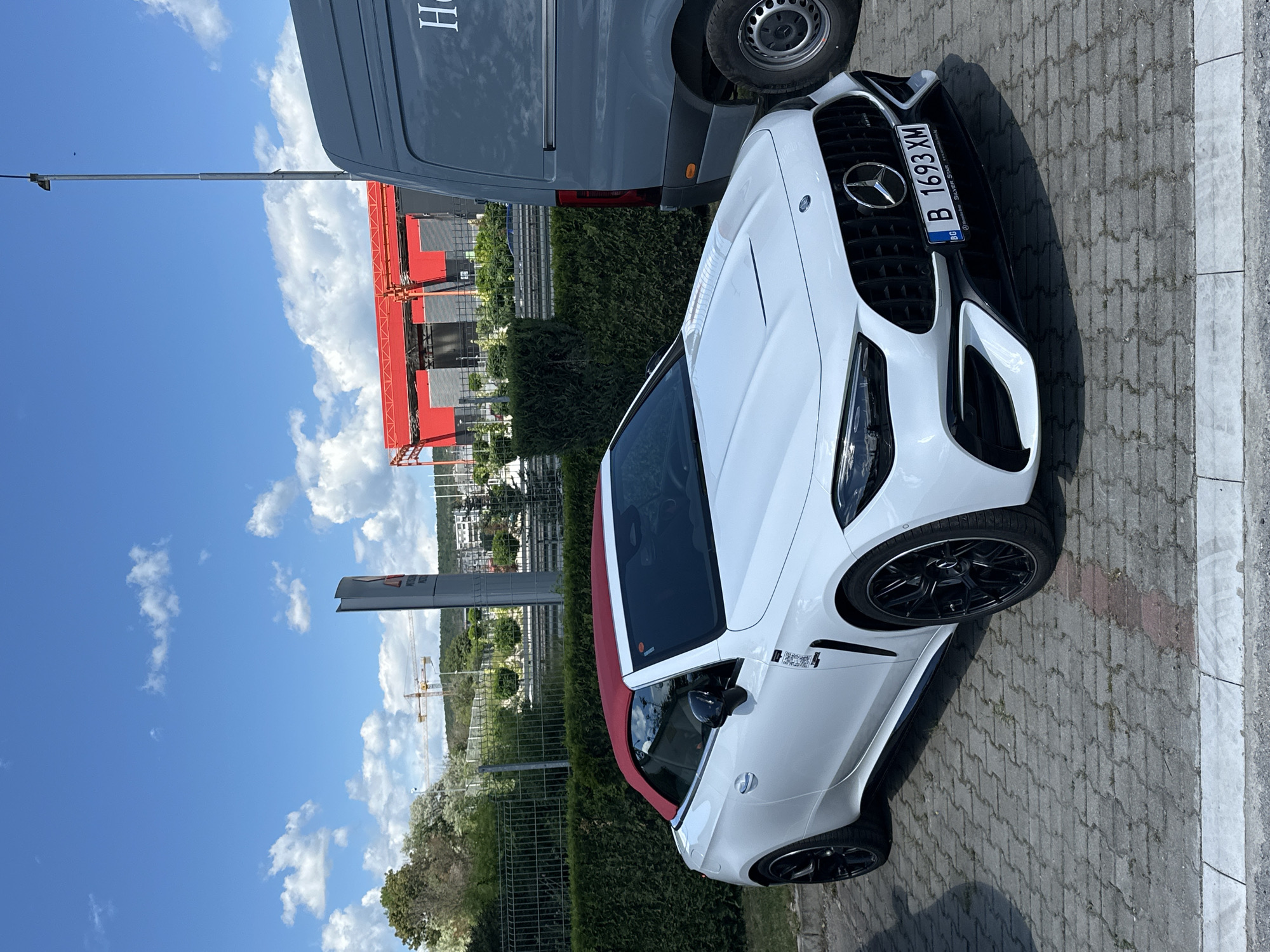 The image shows a white Mercedes-Benz AMG GT convertible parked on a paved surface. The car has a red soft top, black rims, and a Bulgarian license plate "B 1693 XM". Adjacent to the car is a gray Mercedes-Benz van. In the background, there's greenery, buildings, a sign of a business establishment, and a construction crane. The sky is blue with scattered clouds. The setting appears to be an urban environment, likely in the city of Varna, Bulgaria, based on the license plate.