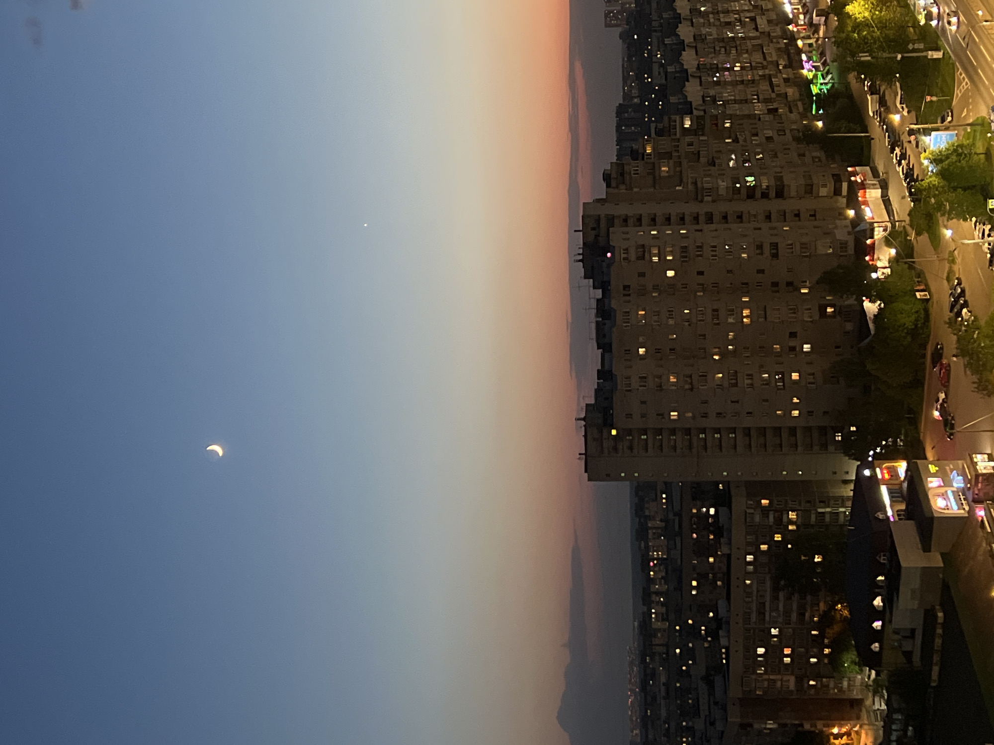 The image depicts a cityscape at dusk, likely taken from a high vantage point in Saint Petersburg, Russia. The sky transitions from a blue on the left to orange on the right. A crescent moon is visible in the blue portion of the sky. A bright dot possibly a star or planet is also visible. The right side of the image shows densely populated urban area, with multiple high-rise buildings. Streetlights and lights from apartments illuminate streets and buildings. Numerous cars and trees are present along the roadside. There are no visible human figures. The overall scene indicates the transition from day to night in a dense urban environment.
