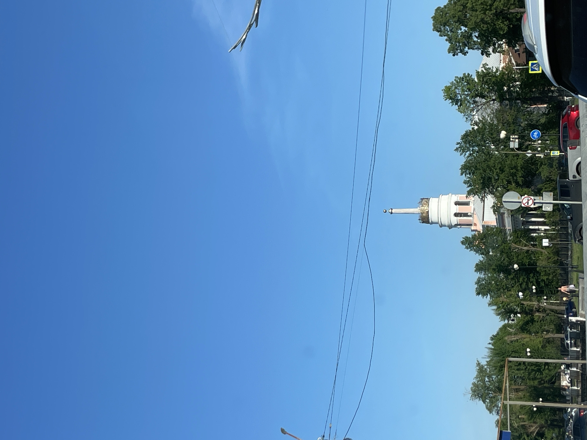 The image depicts a view in Izhevsk, Russia, featuring a tall, multi-tiered building with a spire, likely an architectural landmark. The building is colored white and pink. There are trees on the right-hand side of the frame. A streetscape with streetlights, road signs, and parked vehicles is visible alongside the trees. Overhead electrical wires run across the sky. The sky itself is clear and blue. The photograph appears to have been taken from a vehicle.