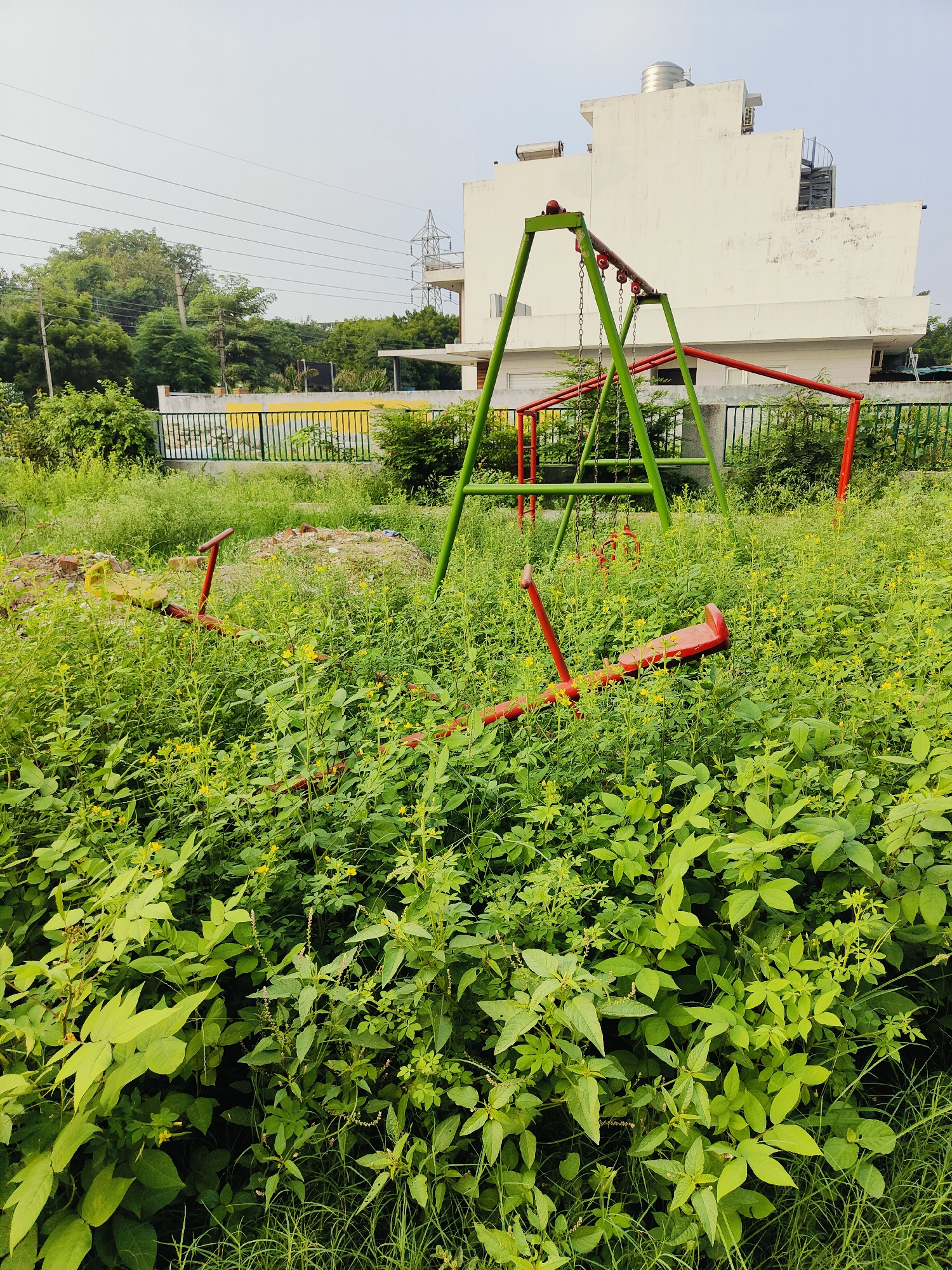 The image depicts an overgrown playground with a swing set and a broken seesaw, situated in Gurgaon, India. The playground is engulfed by lush green vegetation, with tall grass and various plants obscuring much of the ground and the play equipment. The swing set, painted in green and red, stands in the background, appearing functional but also somewhat weathered. In the foreground, a red seesaw lies on its side, partially submerged in the dense foliage, indicating disuse and neglect. The surrounding environment includes a fence, a white building with a water tank on its roof, and power lines, suggesting an urban or semi-urban setting. The sky is overcast, contributing to the muted lighting of the scene. There are no people visible in the image.