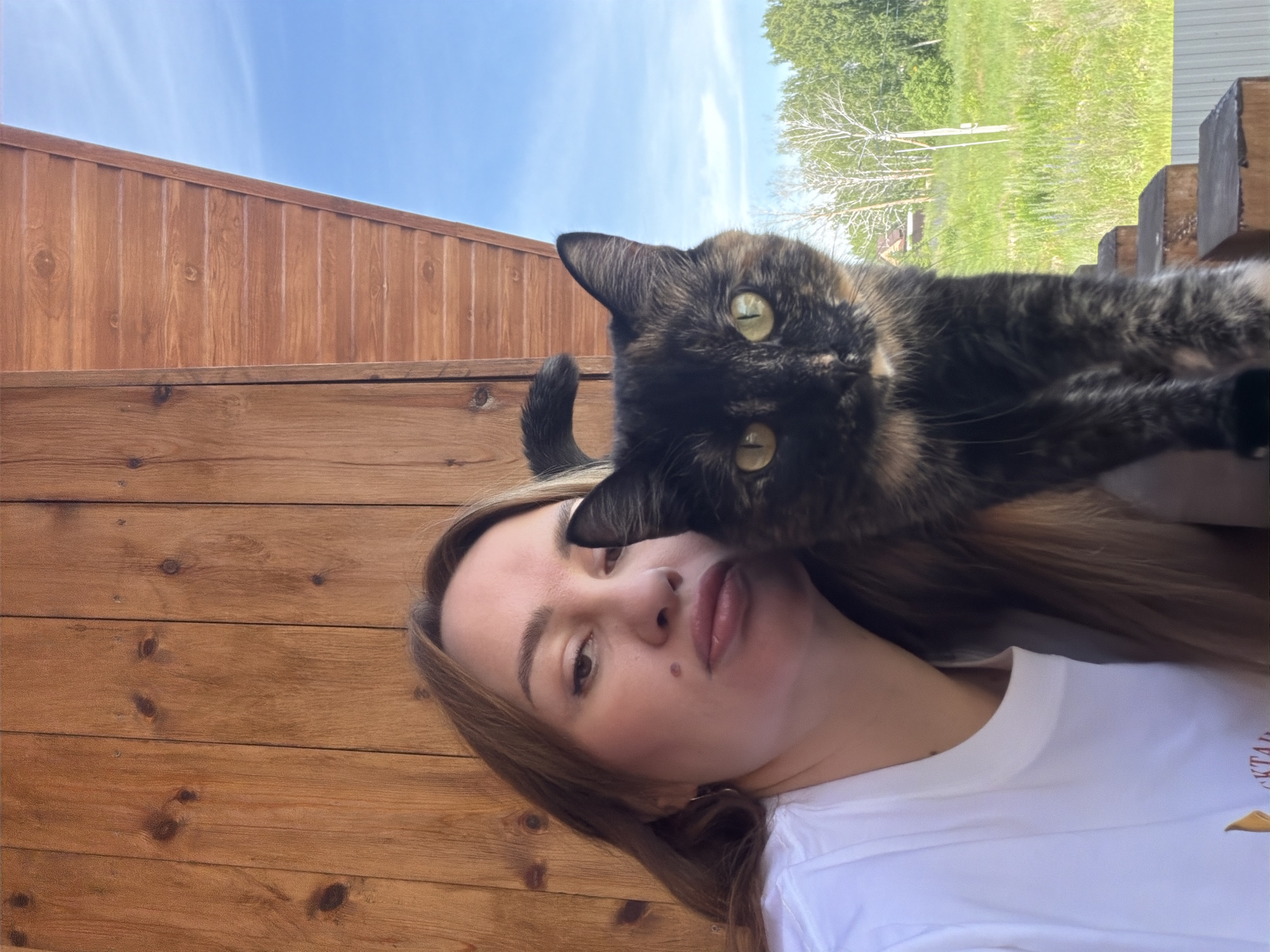 A woman and a tortoiseshell cat are positioned on a wooden porch. The woman, with long, light brown hair, wears a white t-shirt and faces the camera with a slightly puckered expression. The cat, with green eyes, is partially obscuring the woman's face and looks directly at the camera. The background shows a wooden wall and a blue sky with clouds, suggesting an outdoor setting, potentially in or near Malinovka, Russia. The scene appears to be a casual, possibly candid, photograph.