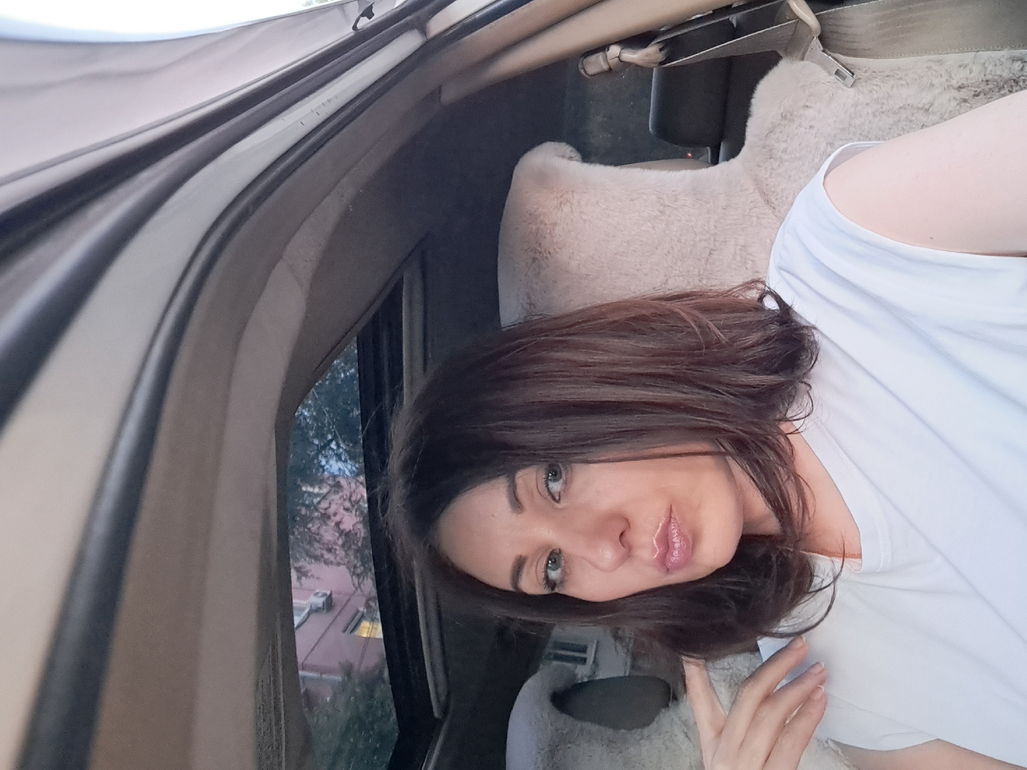A woman with dark brown hair and light green eyes is sitting in the passenger seat of a car with a sunroof open. She is wearing a white t-shirt and her hand is resting on her cheek. Outside the car, a building with pink walls and white windows is visible, along with trees and the sky. The interior of the car is visible, including sheepskin seat covers. The image was taken in Irkutsk, Russia.