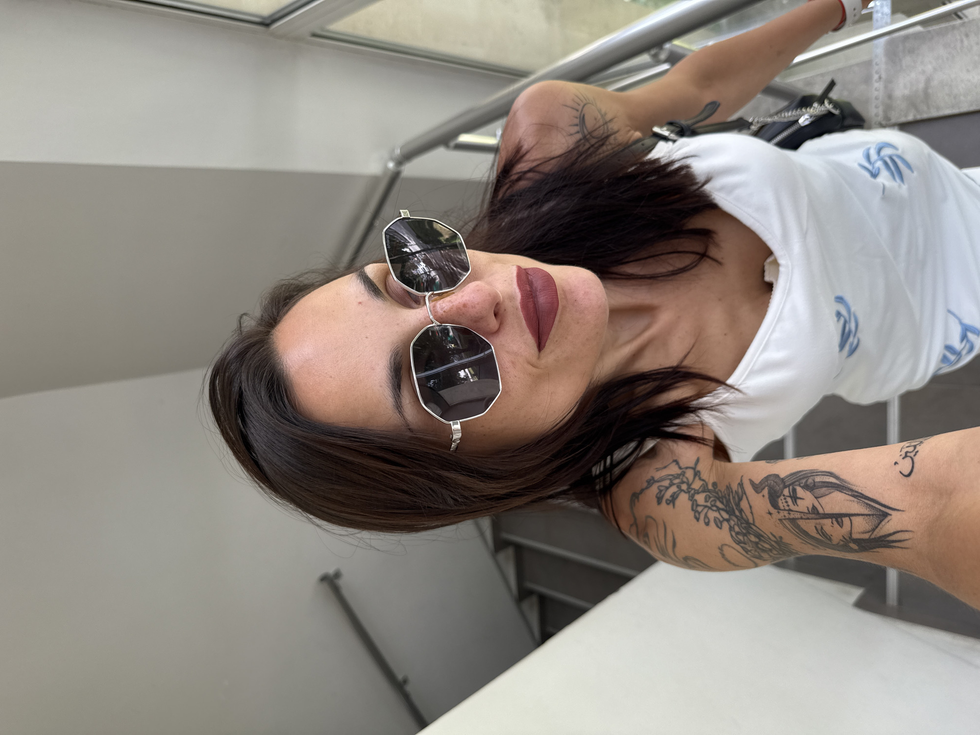 A woman with dark hair, wearing sunglasses and a white dress with blue designs, stands on a staircase. She has tattoos on her arm. The staircase has metal railings and appears to be indoors, possibly in a building in Pleven, Bulgaria.