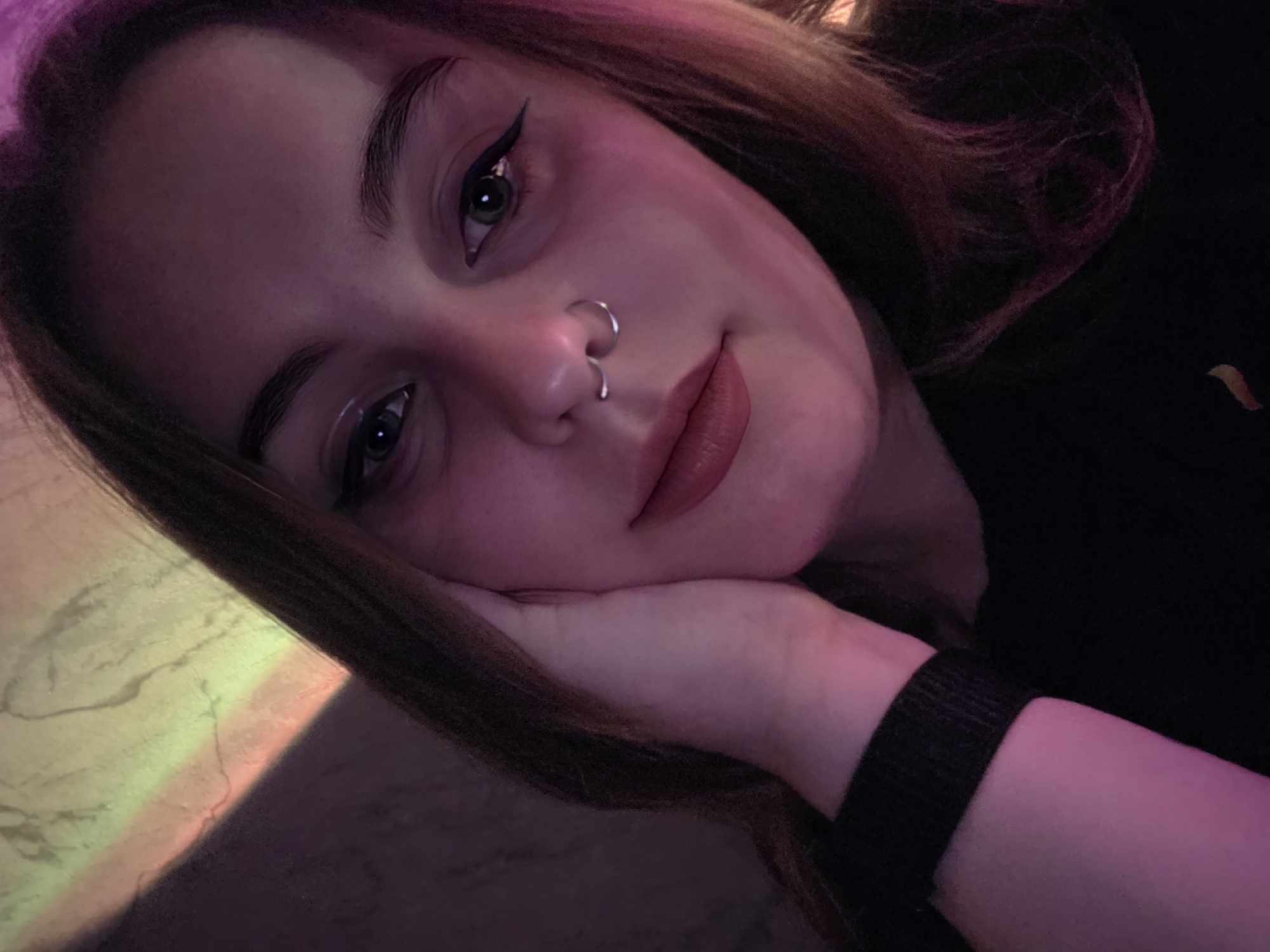 The image is a close-up, overhead shot of a young person resting their head on their hand. The person has brown hair with some pink streaks, makeup including winged eyeliner, a nose ring, and lip gloss. Their skin is illuminated by colored lighting, casting a purple and pink glow on their face and neck. A rainbow-colored light pattern is visible on a textured surface to the left of the person. The person's right arm is visible, with a black band around their wrist. The overall mood is relaxed and possibly melancholic, with the subject's gaze directed slightly away from the camera. The background is dark and nondescript. There is no visible text in the image. The time of day and specific location are not discernible.